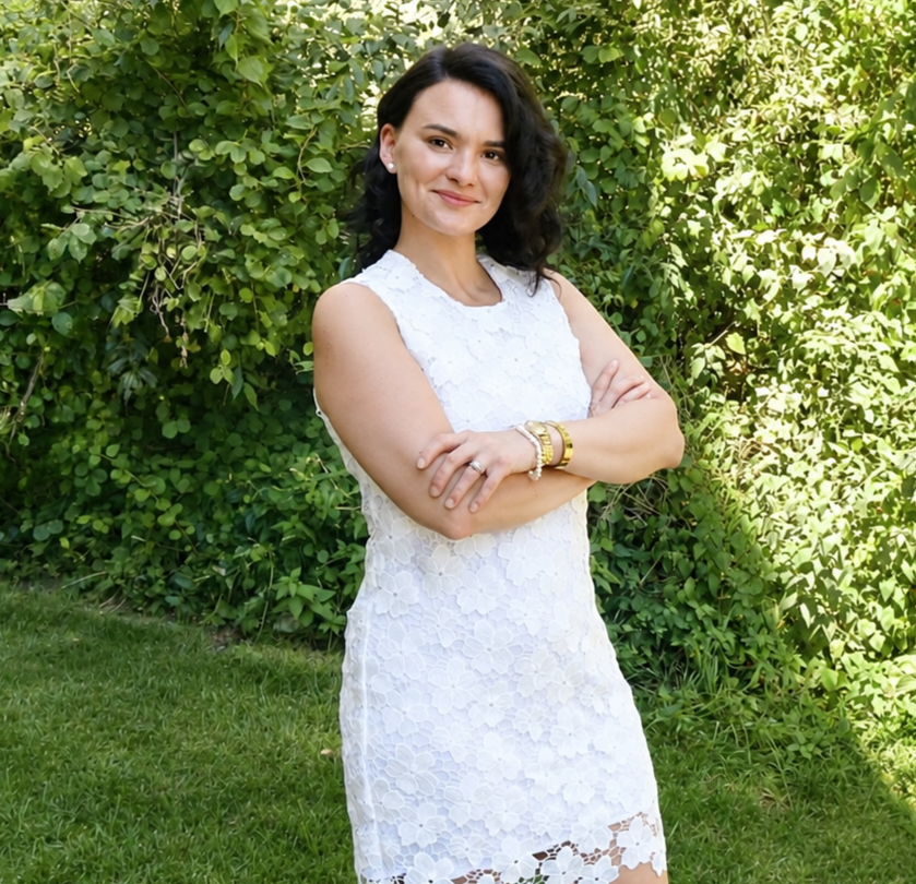 A woman with dark, curly hair standing outdoors in front of green foliage, wearing a sleeveless white lace dress and gold bracelets, smiling with arms crossed.