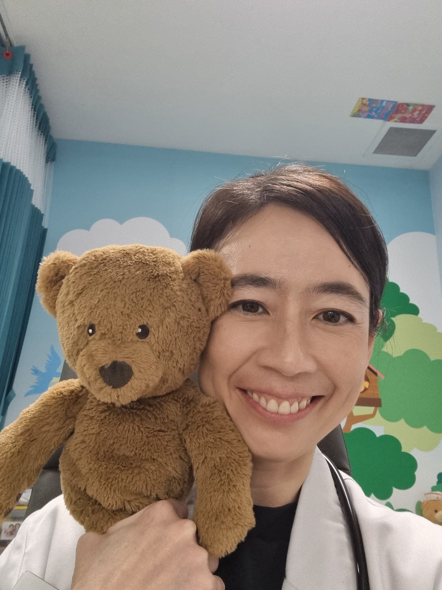 A woman is smiling and holding a brown teddy bear close to her face. She is wearing a white jacket and a black shirt, with a stethoscope around her neck. The background has blue walls with cloud and tree decorations.