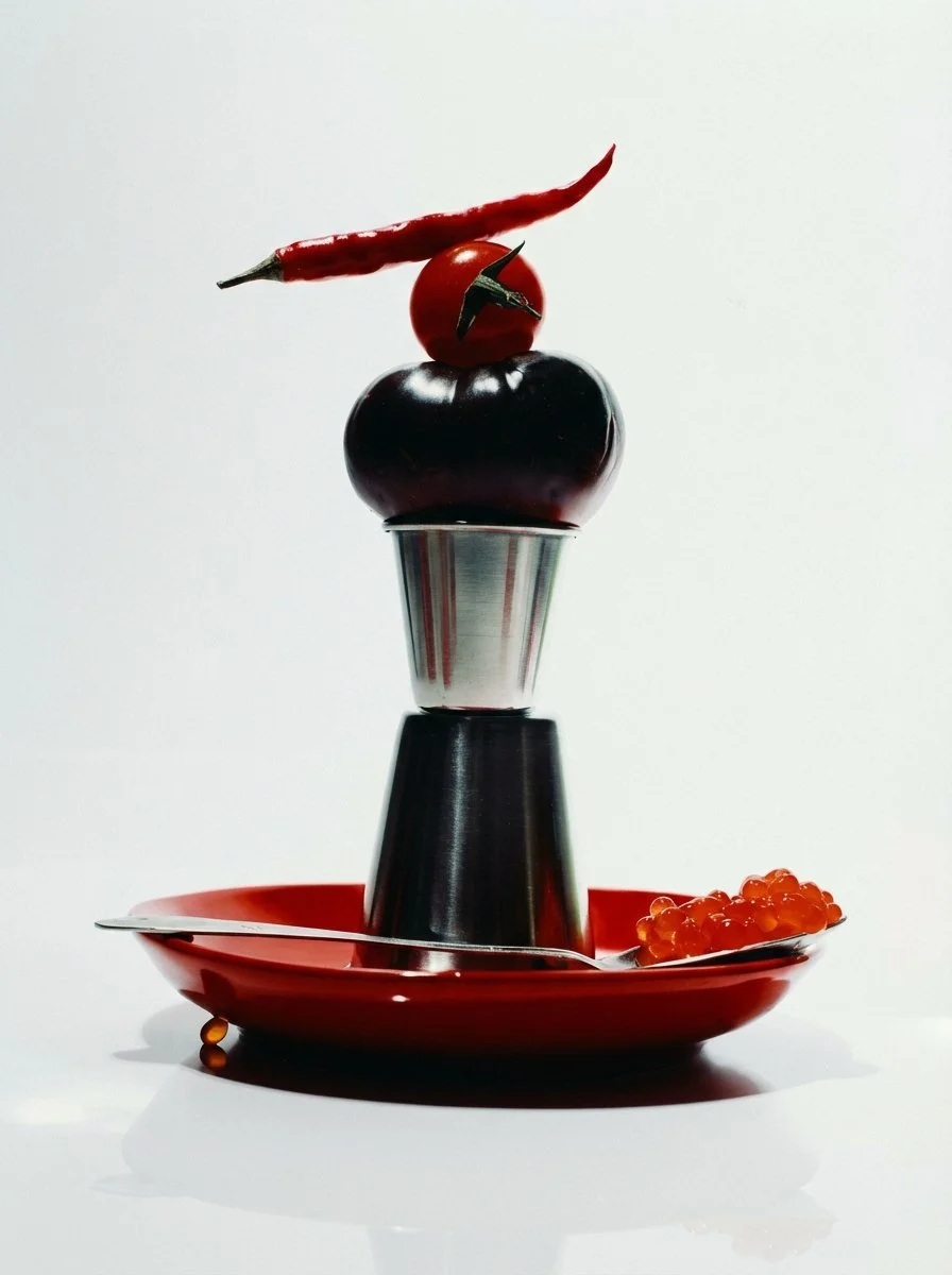 A stack of vegetables including an eggplant, a cherry tomato, and a chili pepper on a metal egg cup, with an orange spoon and fish roe on a red plate against a white background.