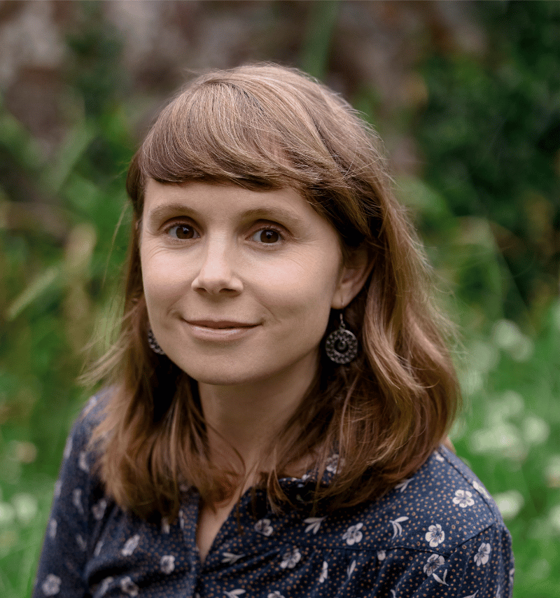 Author Lucy Wood with shoulder-length brown hair and light skin smiling outdoors, wearing a dark blue floral blouse and circular earrings, with a blurred greenery background.