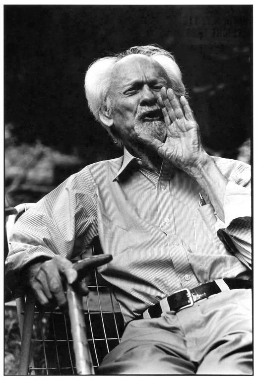 A black and white portrait of Kenneth Burke sitting in a wicker chair, holding a cane. His right hair is held up to his mouth as though to amplify a shout.