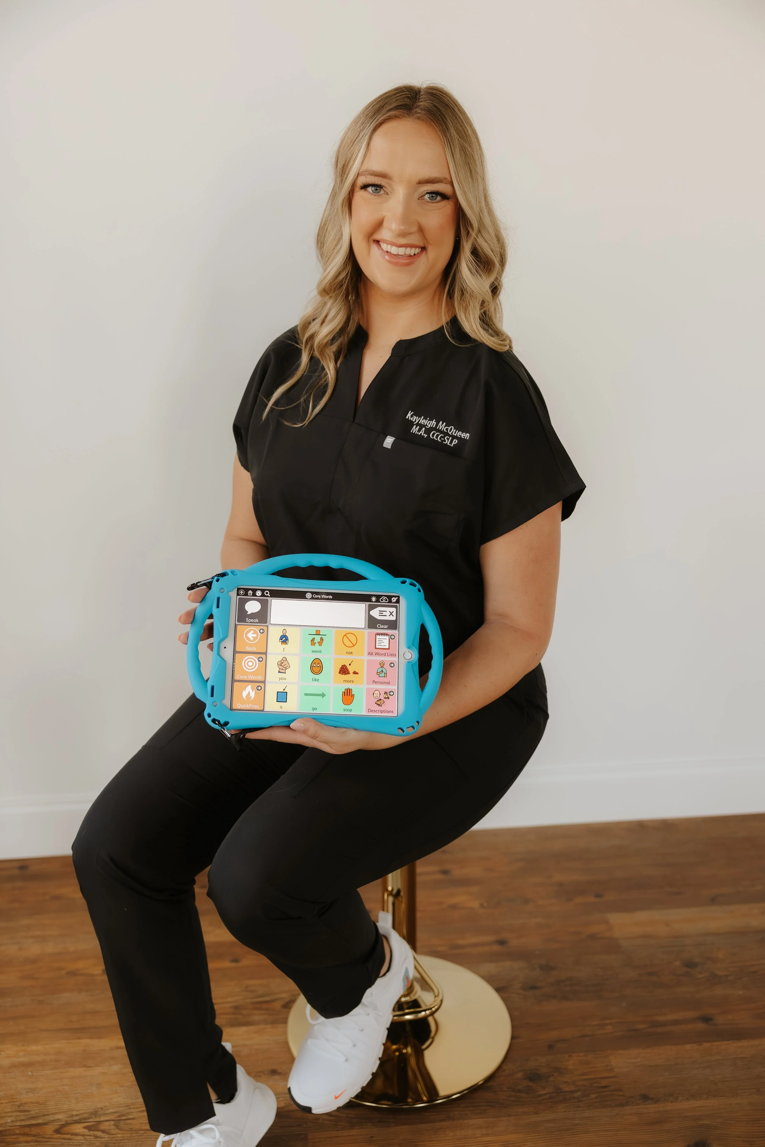 A woman with blonde hair and a black medical uniform sitting on a gold barstool, holding a colorful communication device, in a room with wooden flooring and a plain white wall.