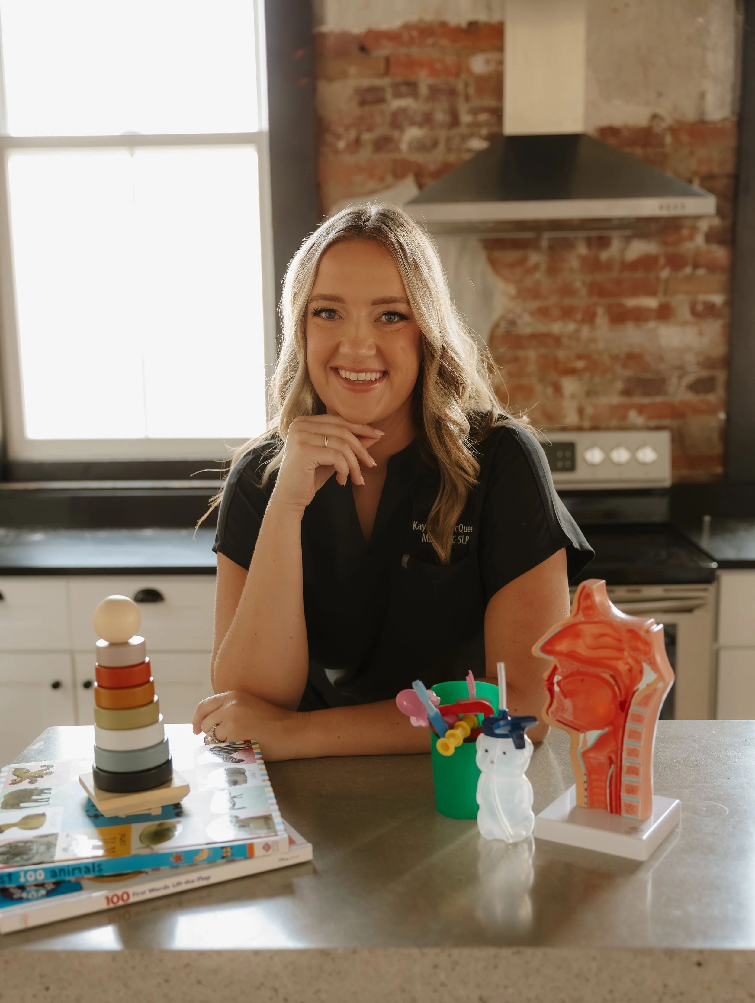 A smiling woman with blonde hair sits at a kitchen counter with educational and creative items, including a children's puzzle, a colorful stacking toy, and a plastic skull and spine model, in a room with exposed brick walls and a large window.