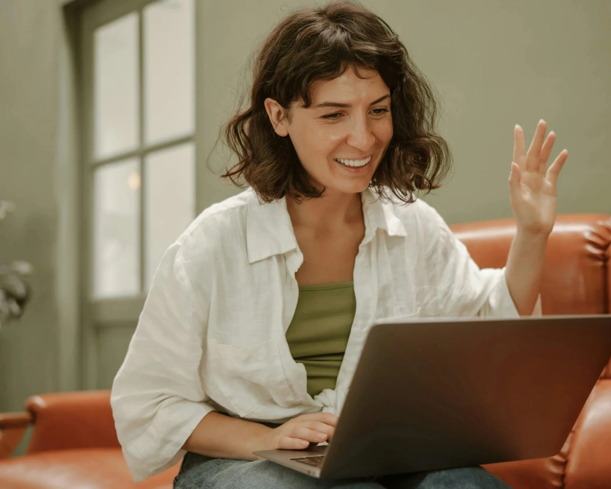 A woman with curly brown hair is attending a virtual therapy session with a psychologist and smiling and waving while sitting on a red chair using a laptop in a room with beige walls and a window in the background.