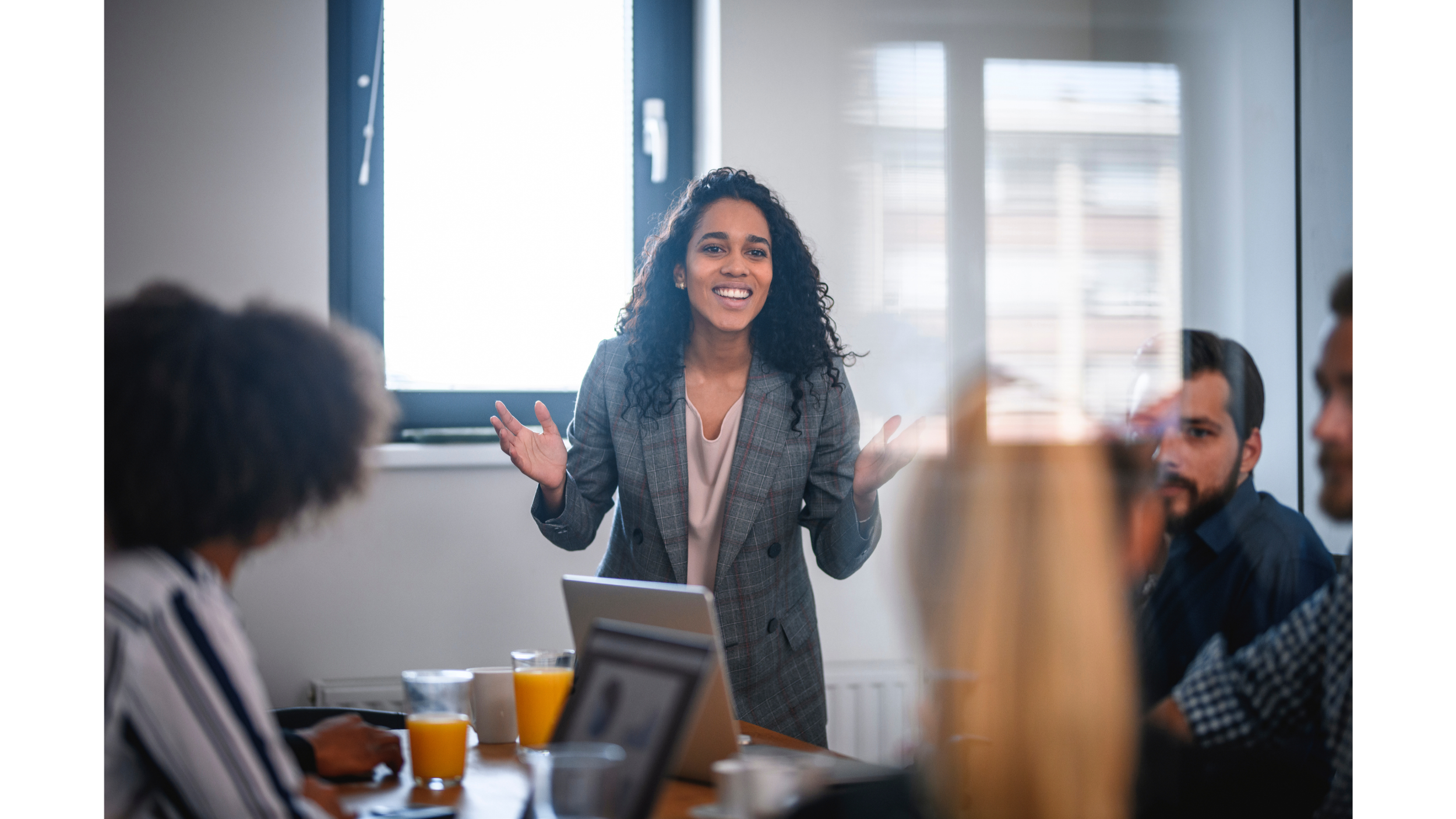 A psychologist with curly black hair, wearing a gray blazer, consulting with a group of people in a conference room during a team training with organization leaders, with some attendees using laptops and glasses of orange juice on the table.