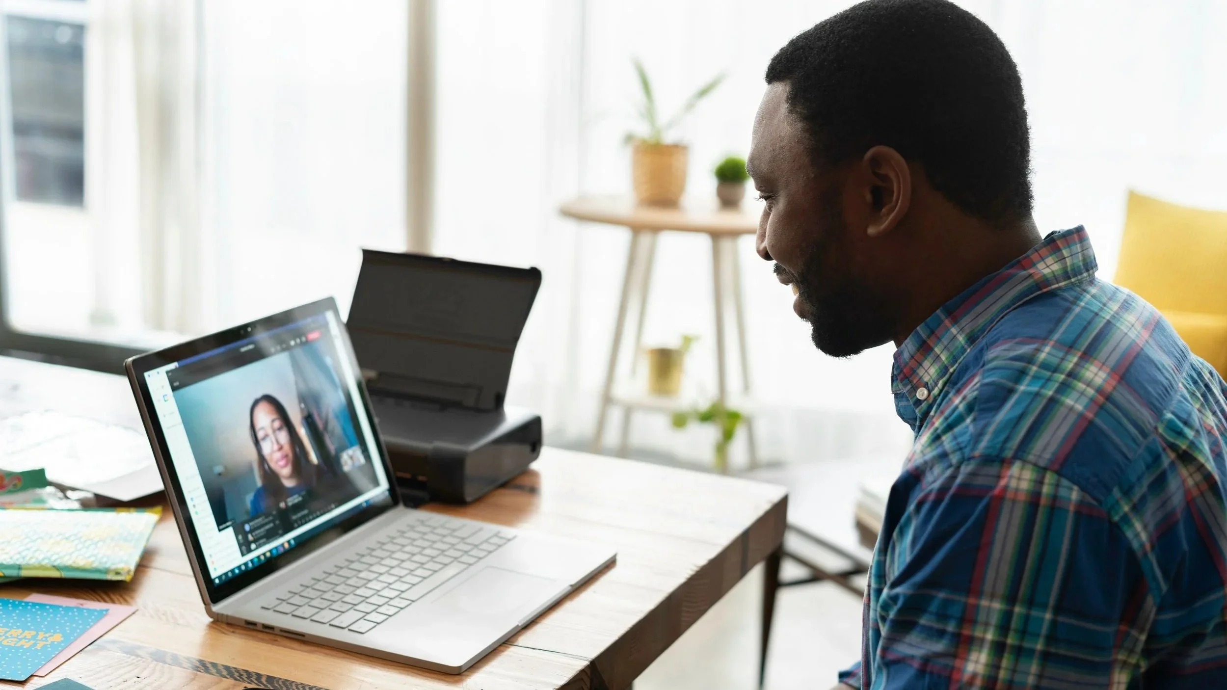 A patient wears a plaid shirt and talks to a psychologist on a telehealth therapy session on a laptop. The man is sitting at a wooden table, and there are notebooks and papers nearby.