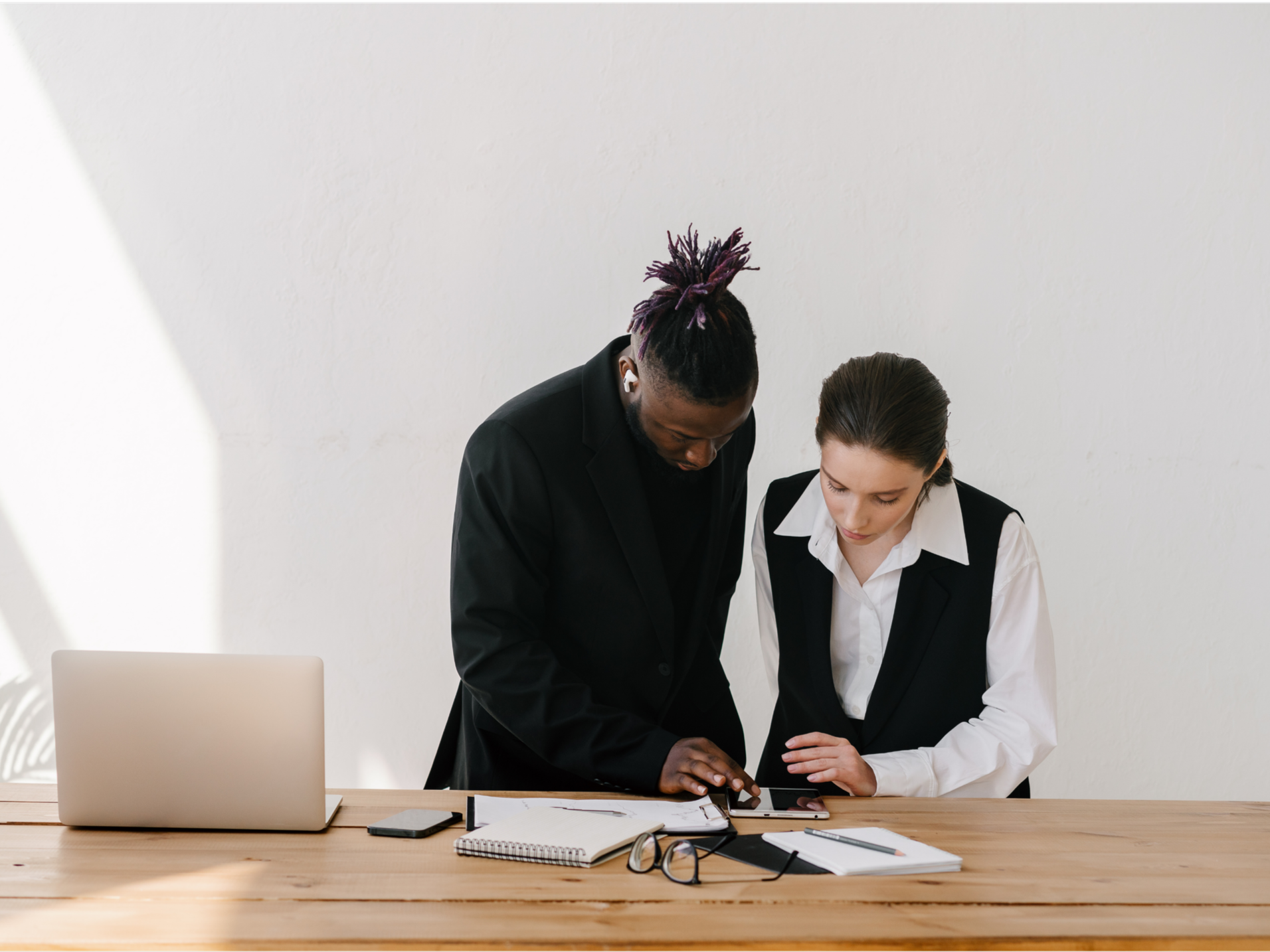 Two healthcare professionals, one man with purple-tipped dreadlocks and a woman with dark hair in a bun, are collaborating at a tablet device in an office setting. The desk has notebooks, glasses, a smartphone, and a laptop.