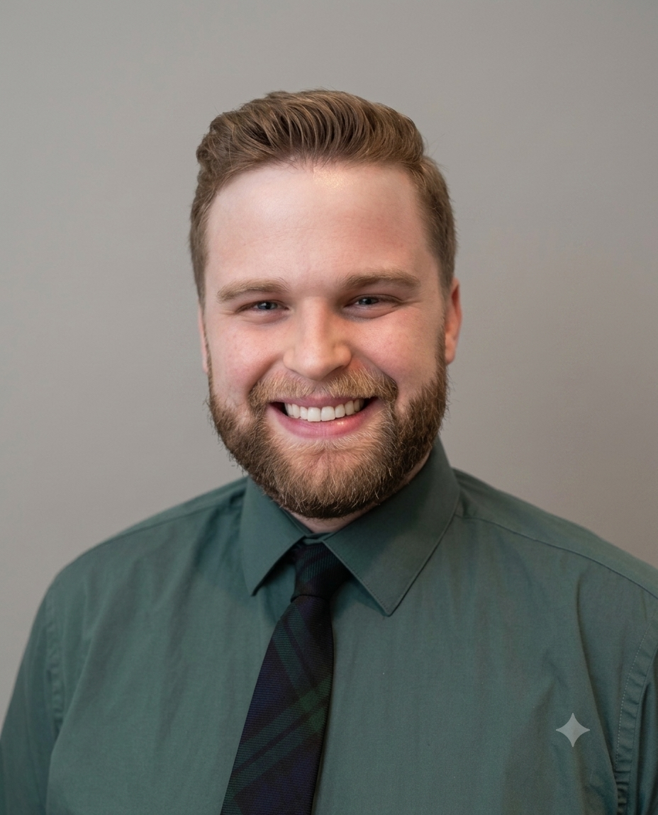 A psychologist and behavioral science consultant with a beard and short hair, smiling, wearing a green dress shirt and a dark green and black patterned tie, against a plain light gray background.