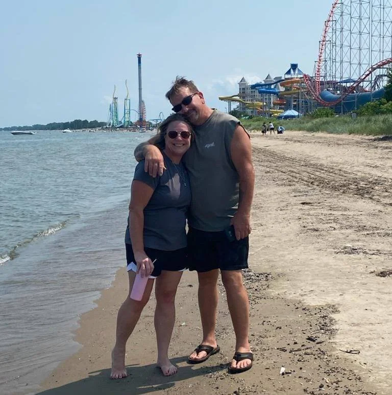 A couple standing on a sandy beach, smiling, with amusement park rides in the background, including a roller coaster and water slides.