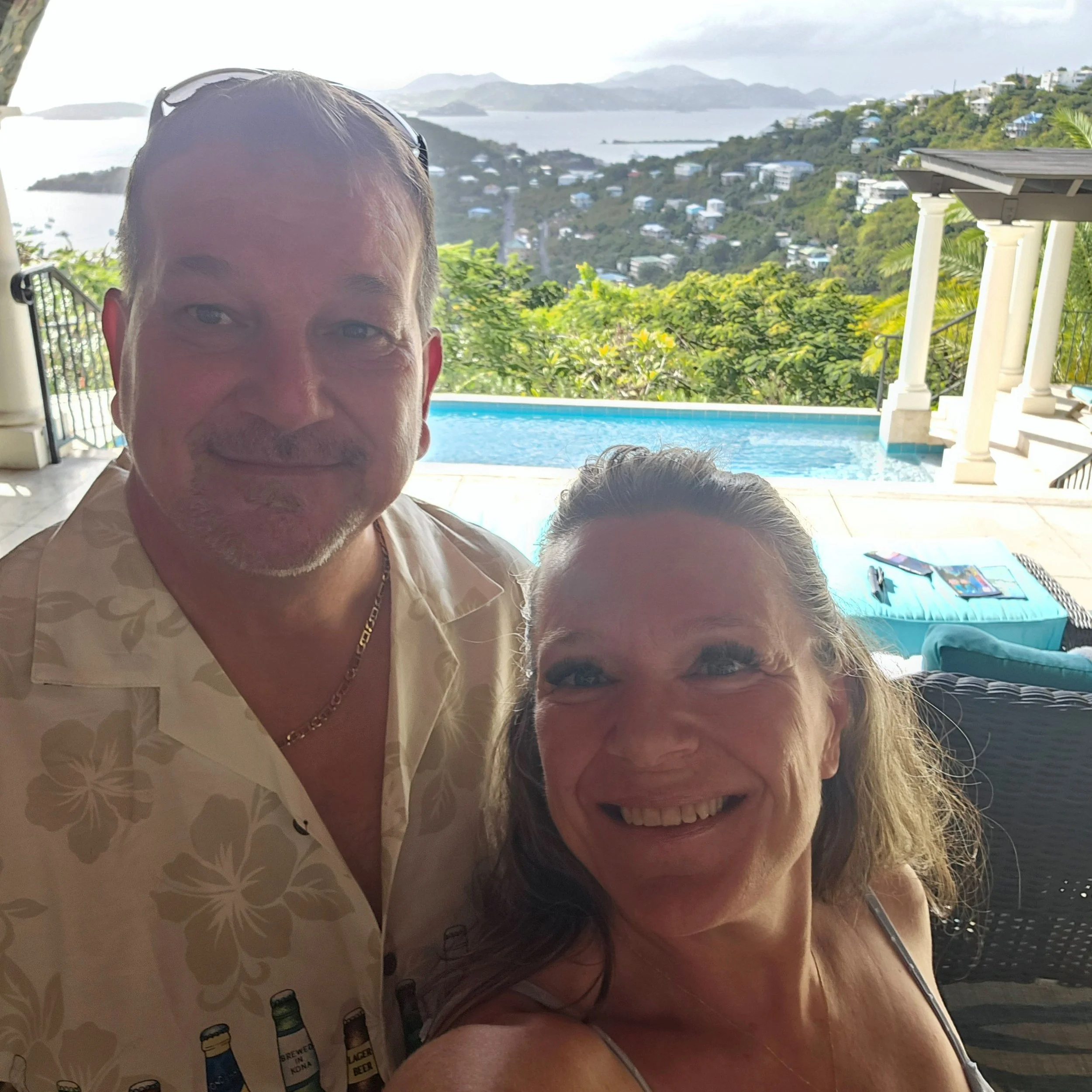 A man and a woman are close together, smiling, with scenic water and green hills in the background. They appear to be outdoors near a swimming pool, with a partly cloudy sky overhead.