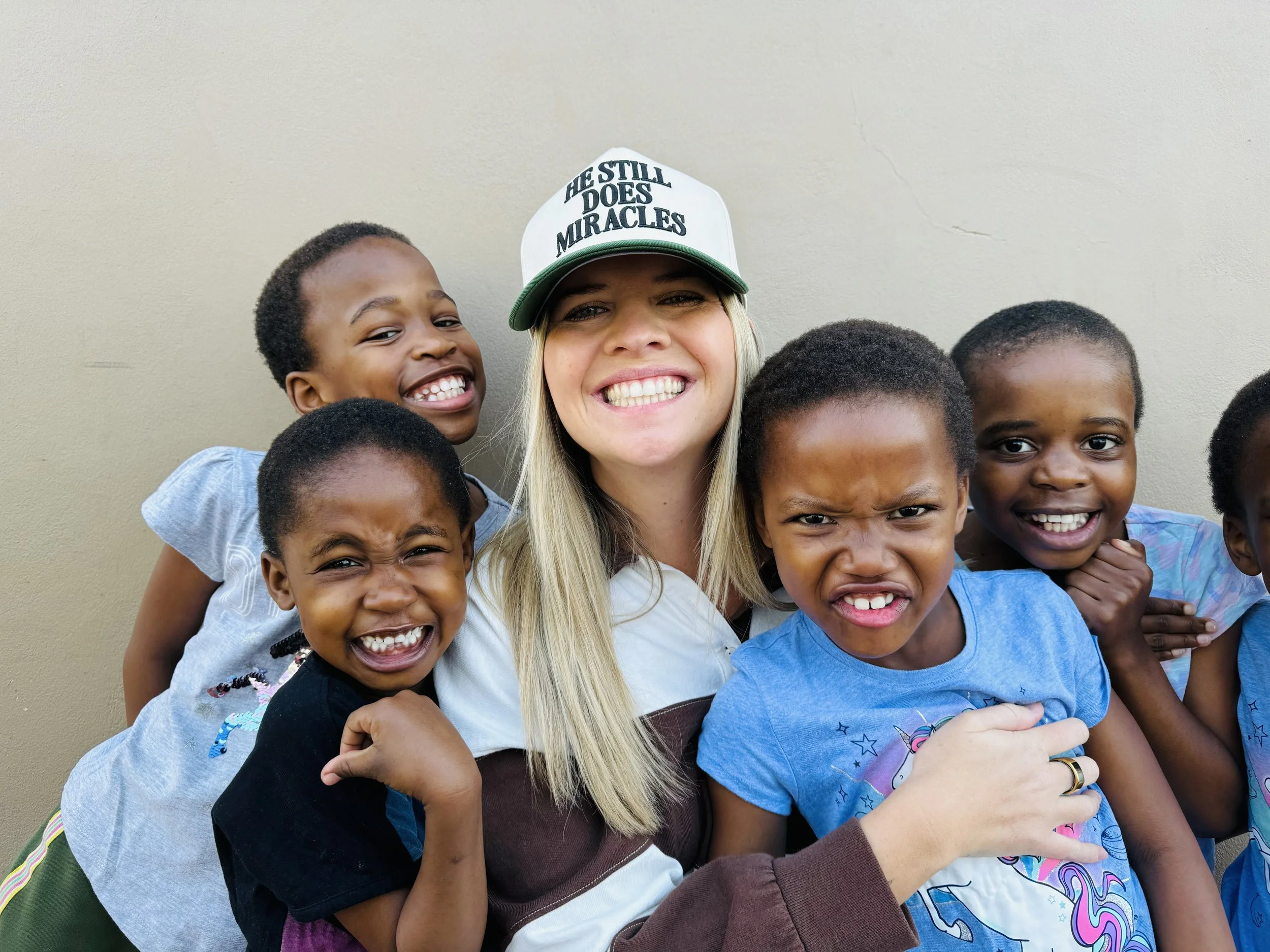 A group of children and a woman smiling and hugging each other against a plain wall. The woman is wearing a baseball cap that reads 'He still does miracles'.