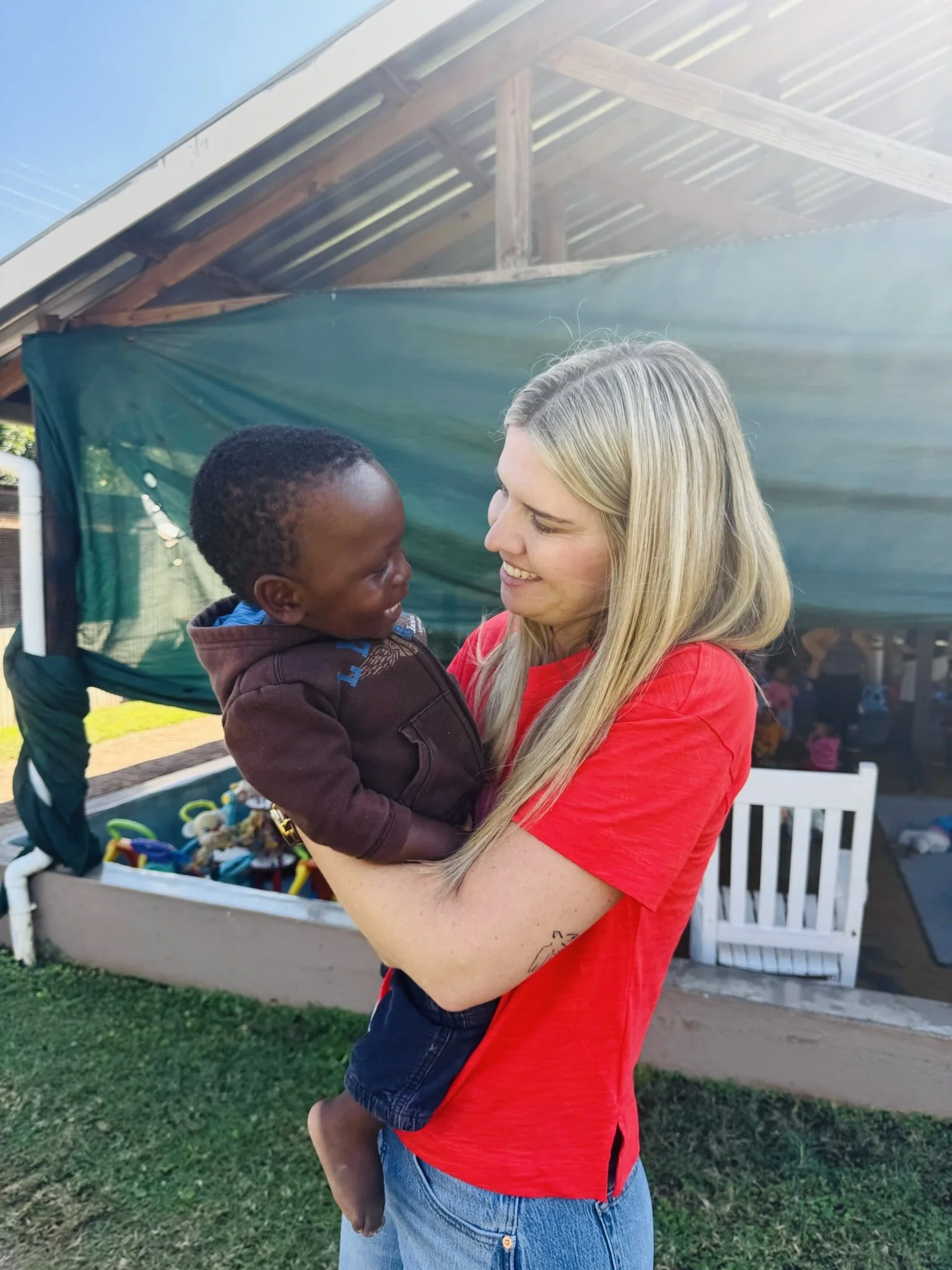 A woman with long blonde hair holding a young boy with short curly hair, while smiling at him outdoors in a backyard with green grass, a white chair, and a canopy tent in the background.