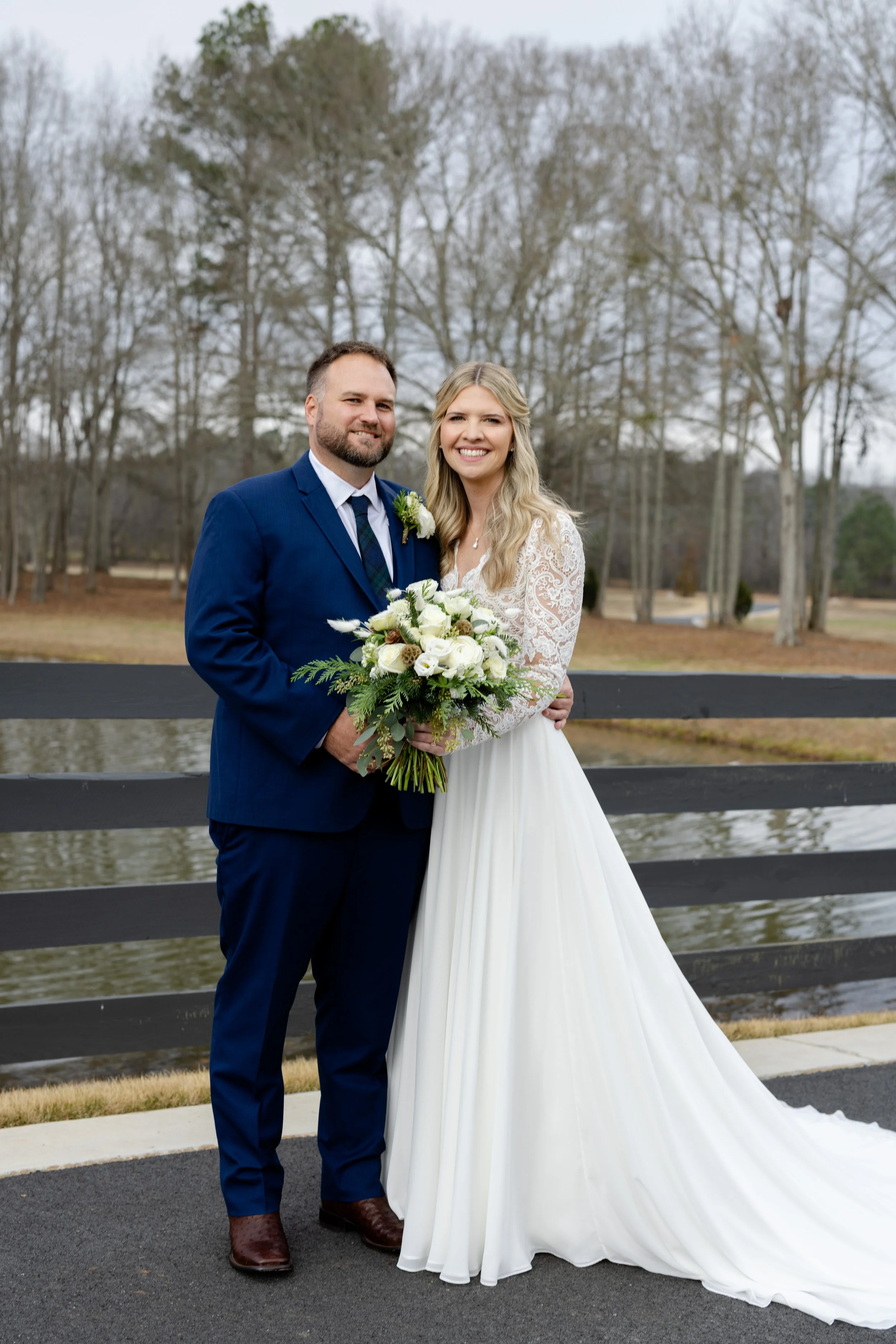 A bride and groom standing outdoors near a lake and trees, smiling. The groom is in a blue suit with a boutonniere, and the bride is in a long white lace wedding dress holding a bouquet of white flowers and greenery.
