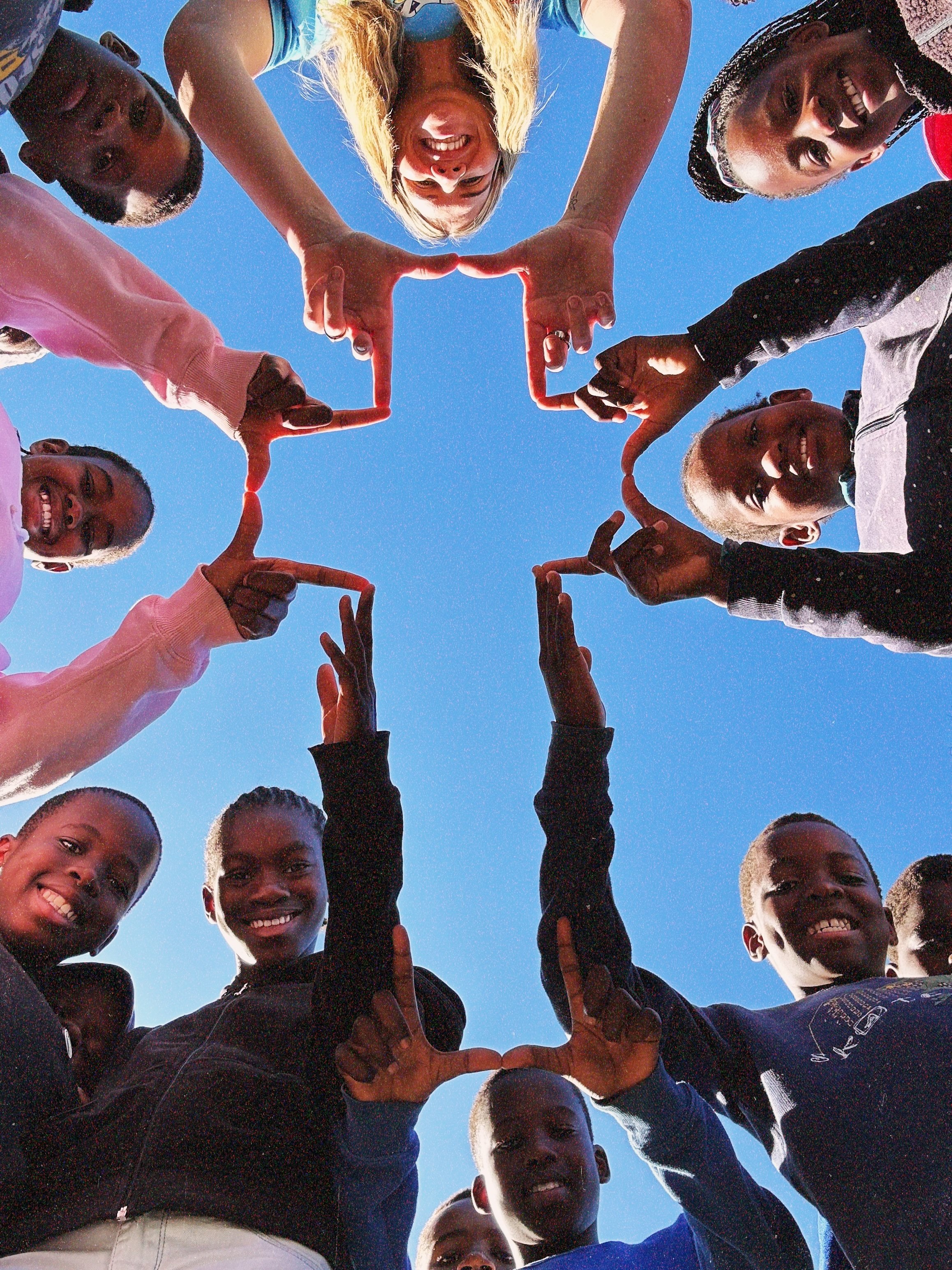 Group of diverse children and a young woman forming a heart shape with their hands while looking down at the camera, smiling, with a clear blue sky in the background.