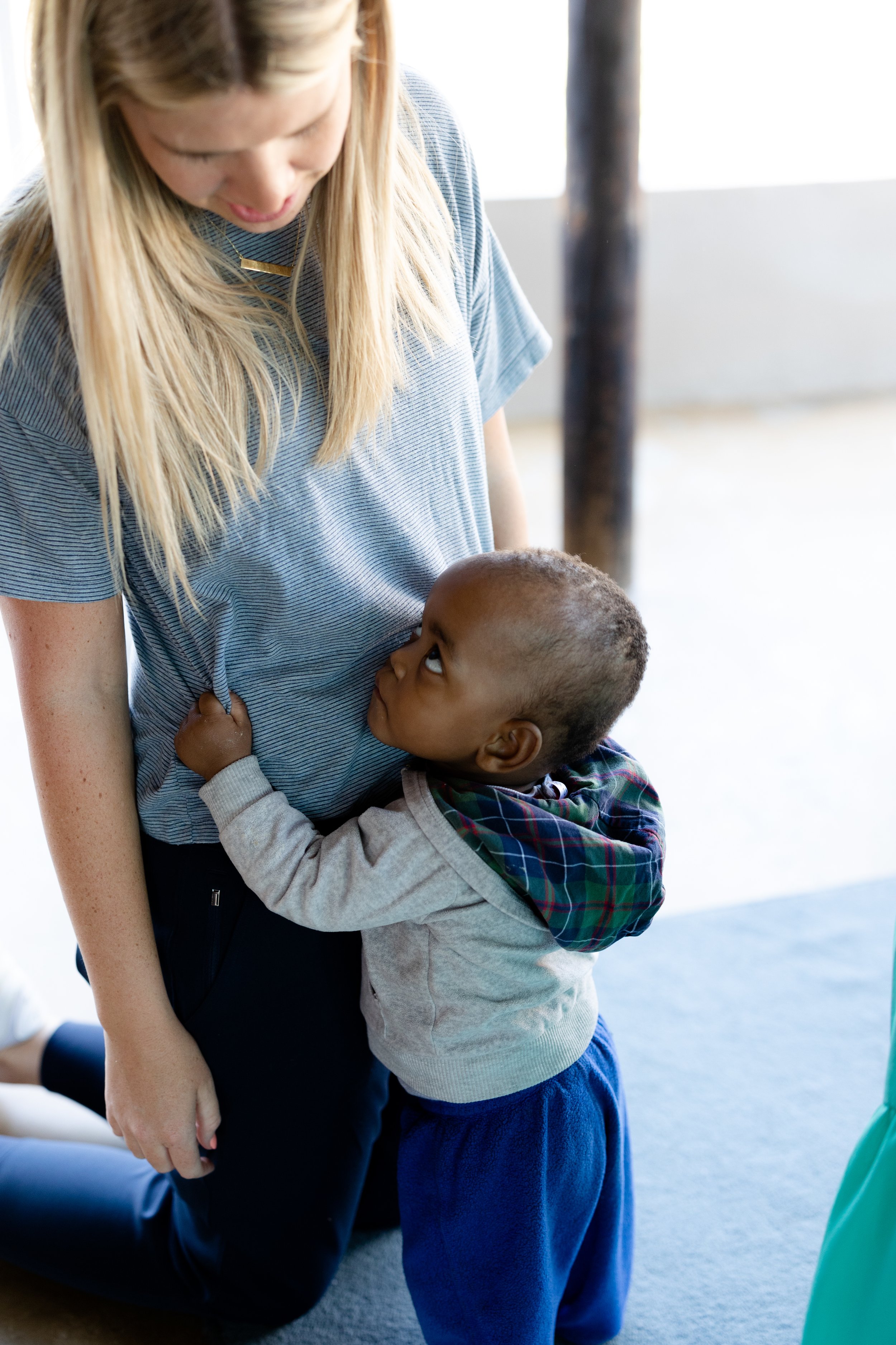 A woman with blonde hair and a striped blue shirt kneels down as a young boy with dark hair and a plaid shirt hugs her around the waist, looking up at her.