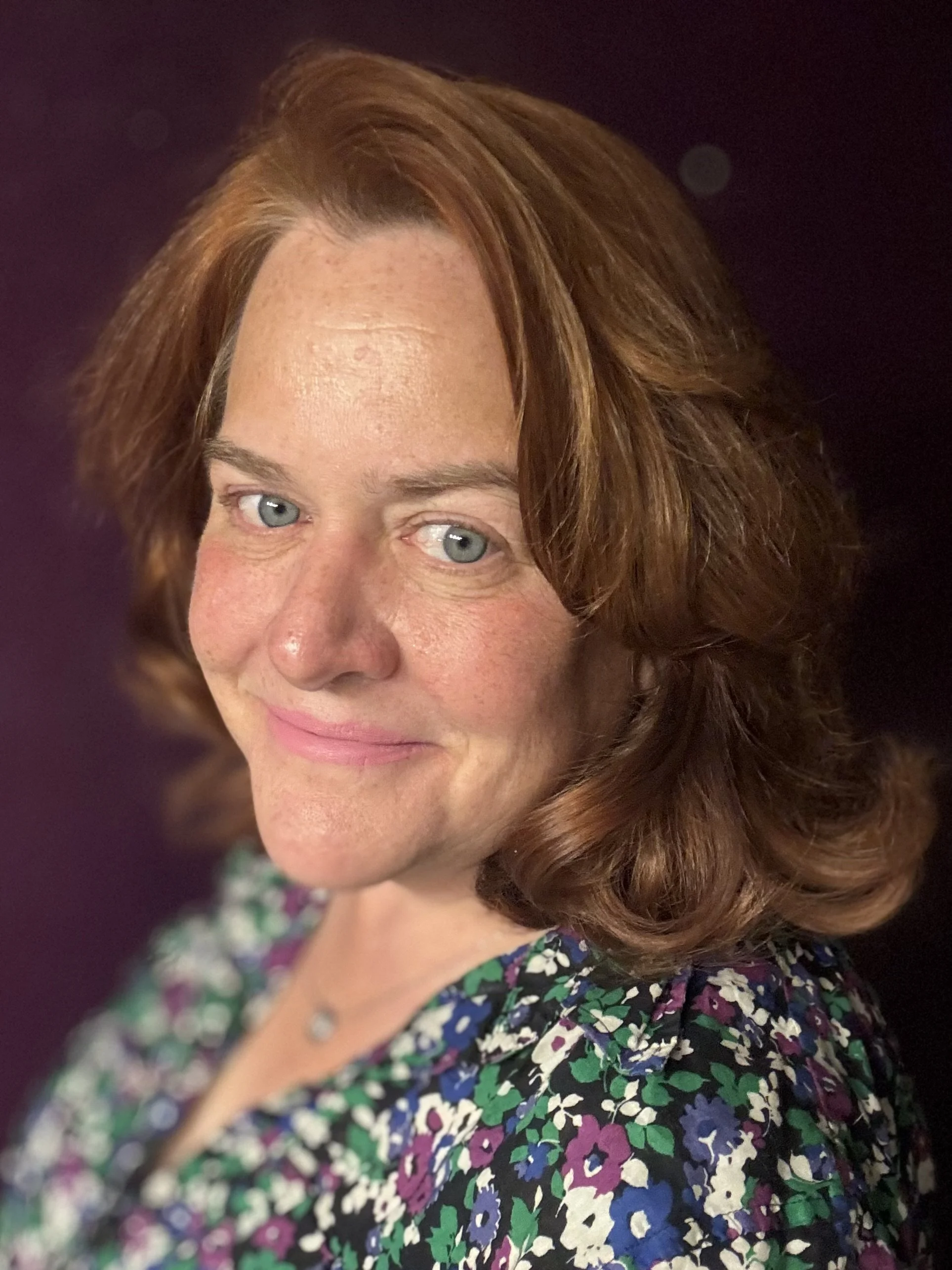 Close-up of a woman with fair skin, red hair, light blue eyes, and wearing a floral dress with blue, purple, green, and white colors, against a dark background.