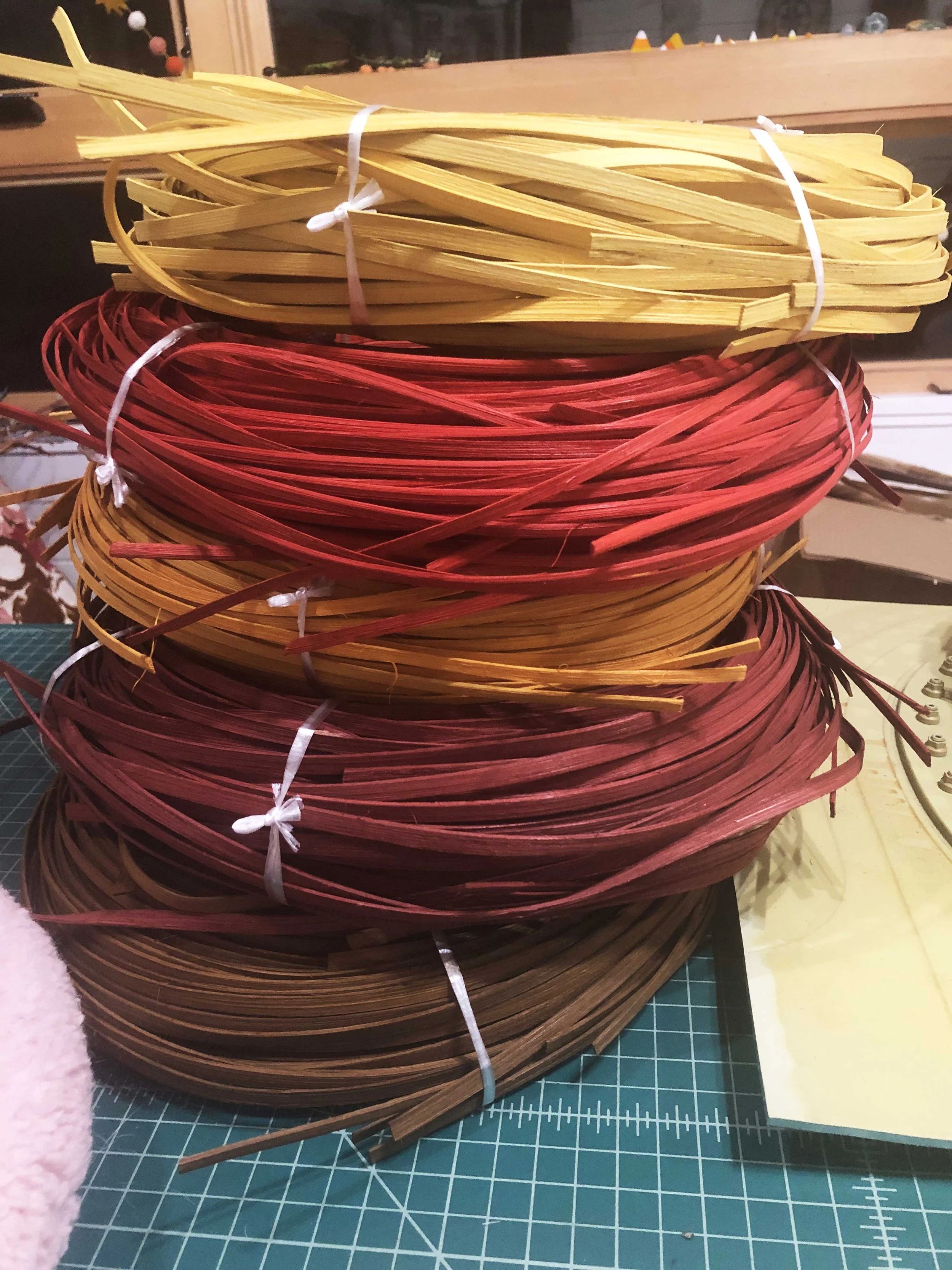 Stacks of colored woven craft material, including yellow, red, orange, and brown, tied with white strings on a green cutting mat with measurement markings.