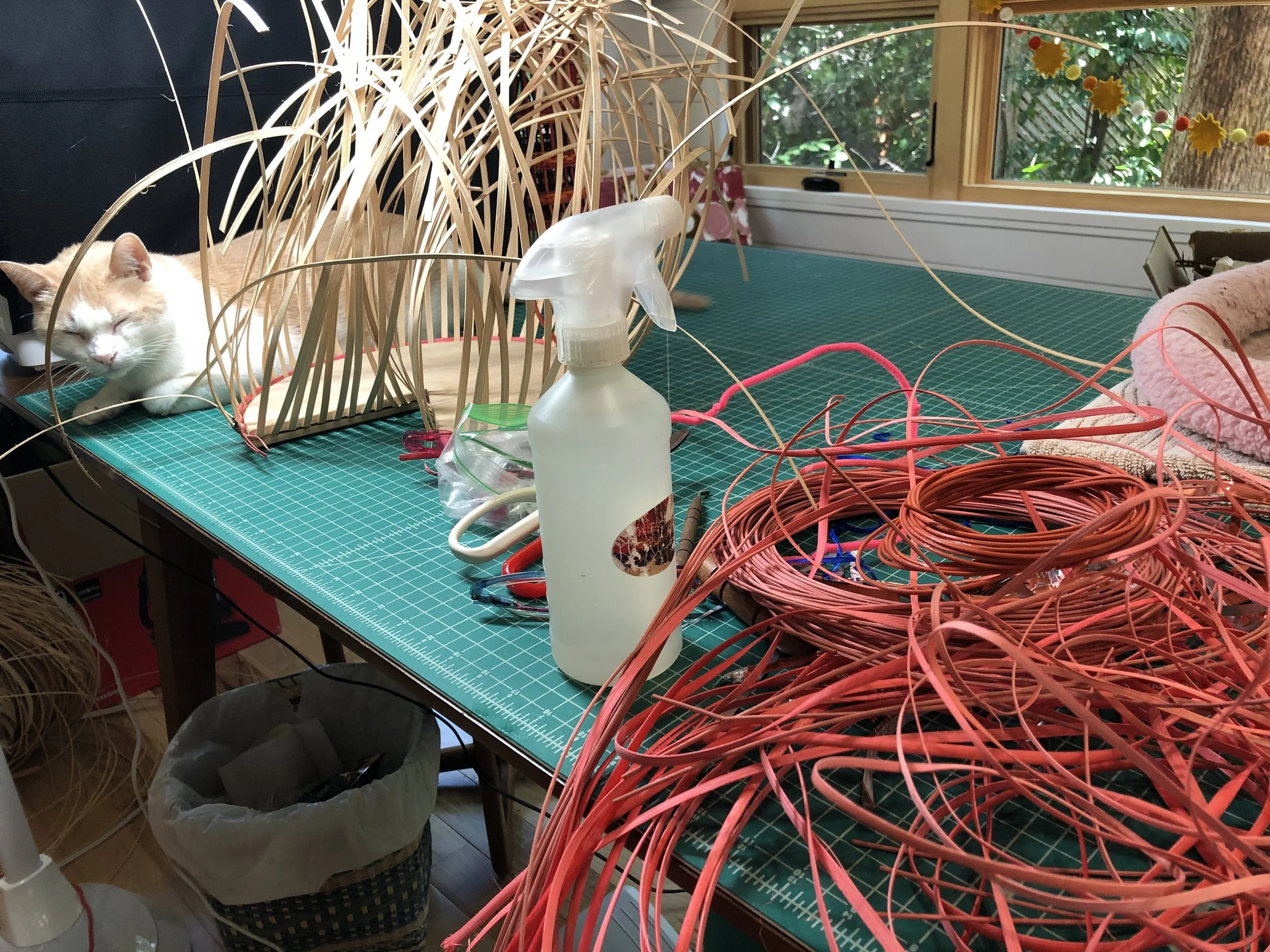 A worktable in a room with large windows showing trees outside. The table has various crafting supplies, including orange and pink wires, tools, and a plastic spray bottle. A tabby cat with orange and white fur is sleeping on the wooden surface, partially hidden behind a woven basket filled with scraps.