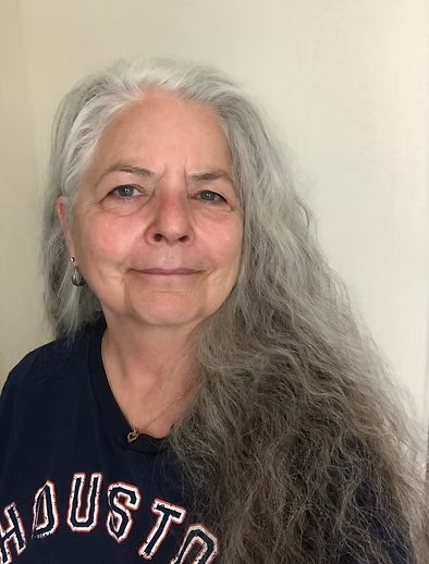 A woman with long, wavy gray hair and light skin, smiling slightly, wearing a black shirt with 'Houston' written on it, standing against a plain light-colored wall.