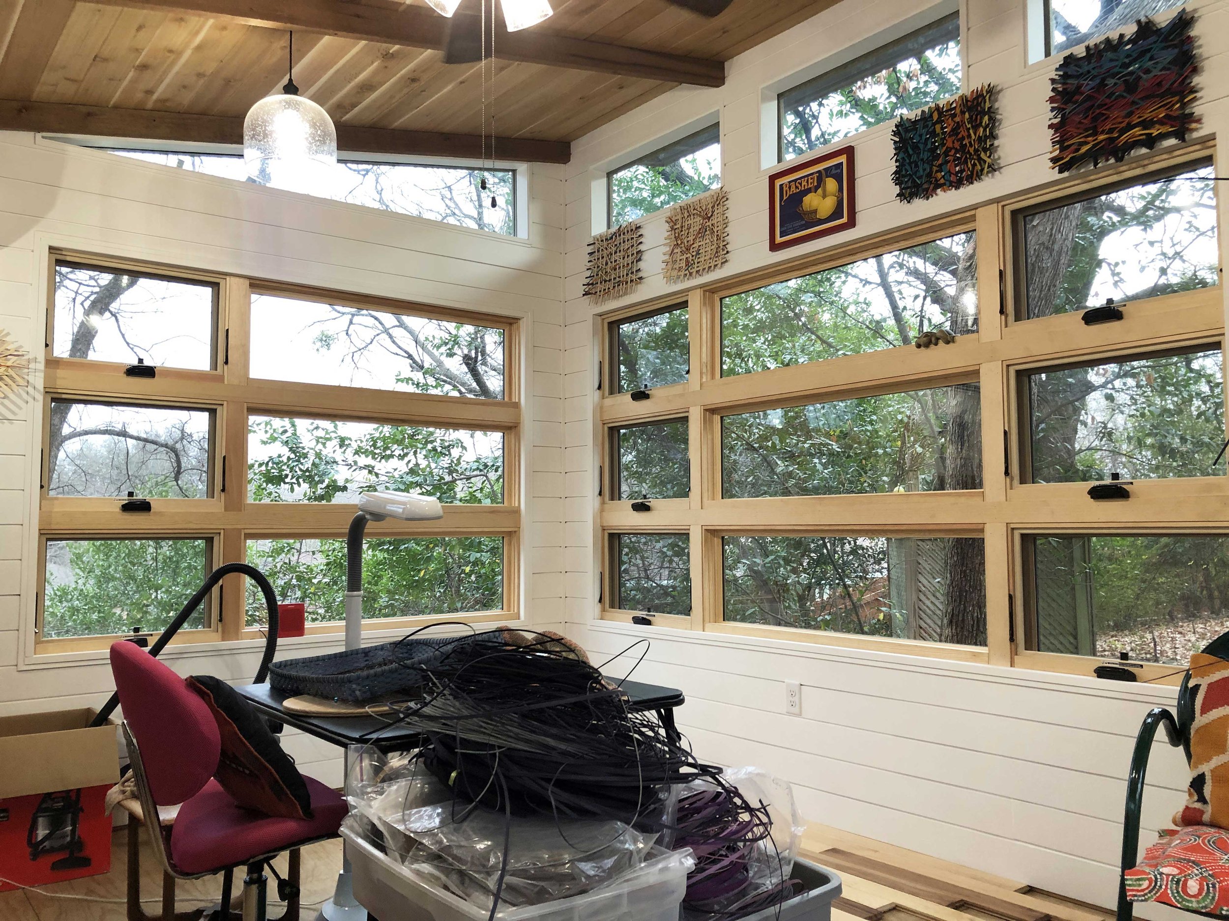 Interior of a room with multiple windows showing trees outside. There is a ceiling fan and hanging light fixtures. The room appears to be under construction with tools, wires, and materials on the table and around.