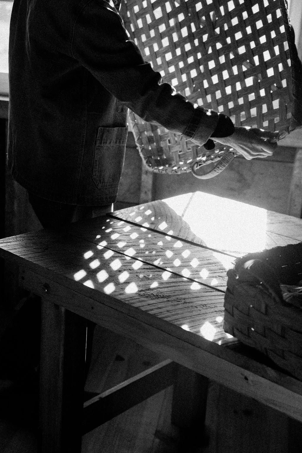 A person in a denim jacket holding a pair of gloves and a drink, standing near a wooden table with a woven basket on it, with sunlight creating a diamond pattern through a lattice structure.