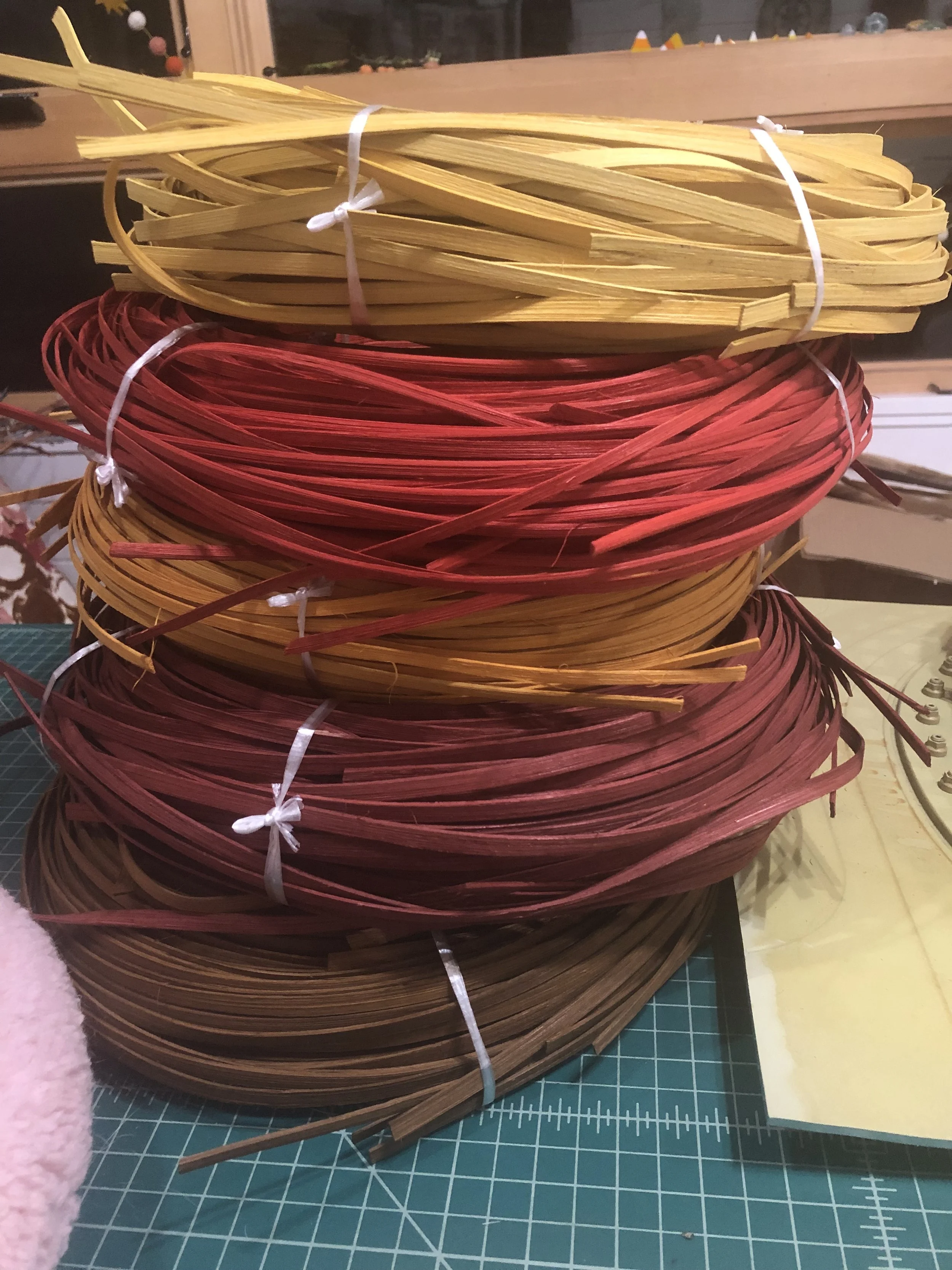 Multiple stacks of woven craft materials in yellow, red, orange, and brown colors, tied with white ribbons, placed on a green cutting mat in a workspace.