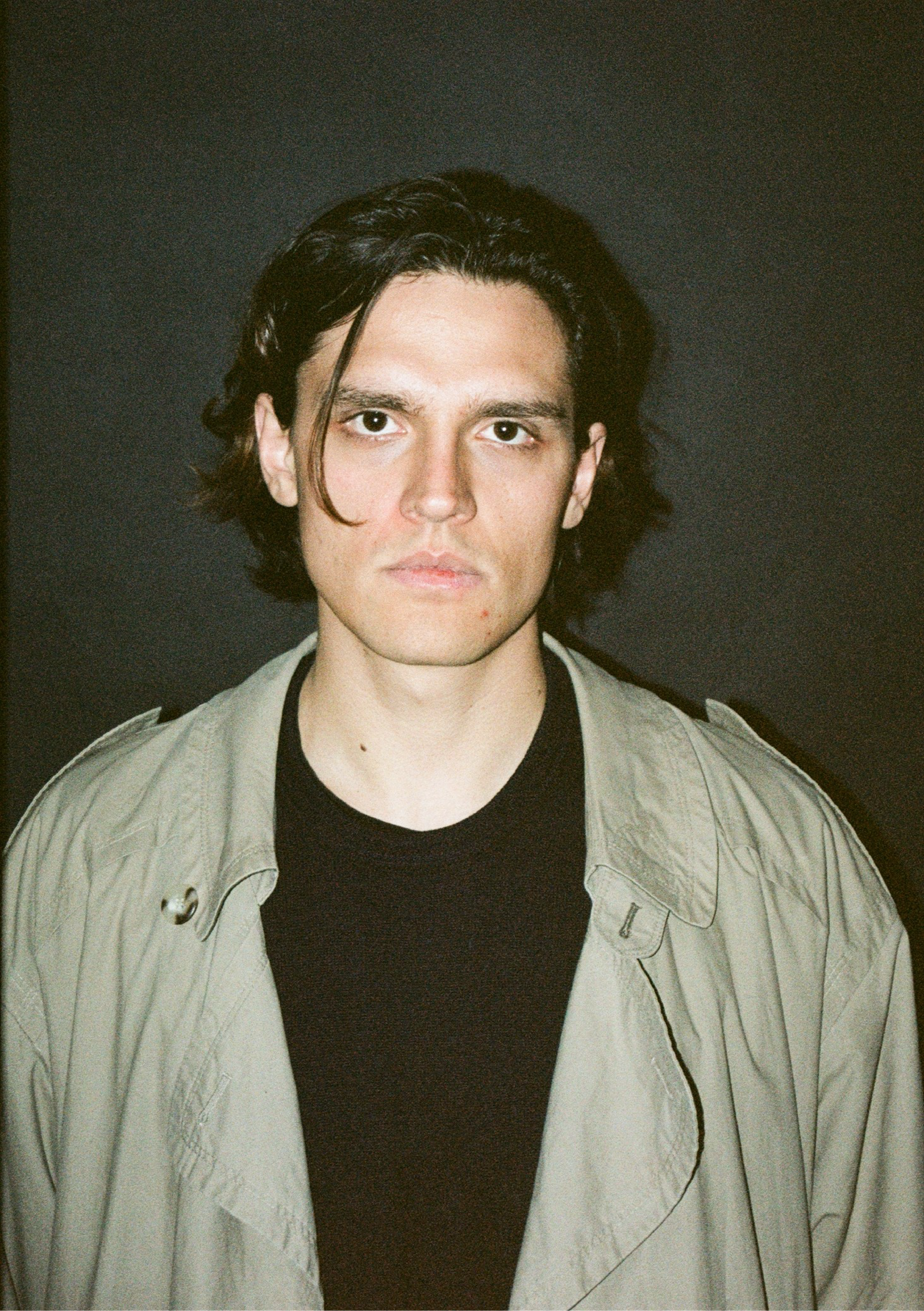 A young man with black hair, wearing a black shirt and a light-colored jacket, standing against a dark background.