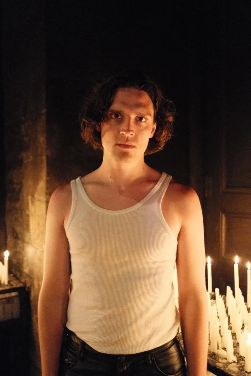 A young man with curly hair in a white tank top standing amidst candlelight in a dark room.