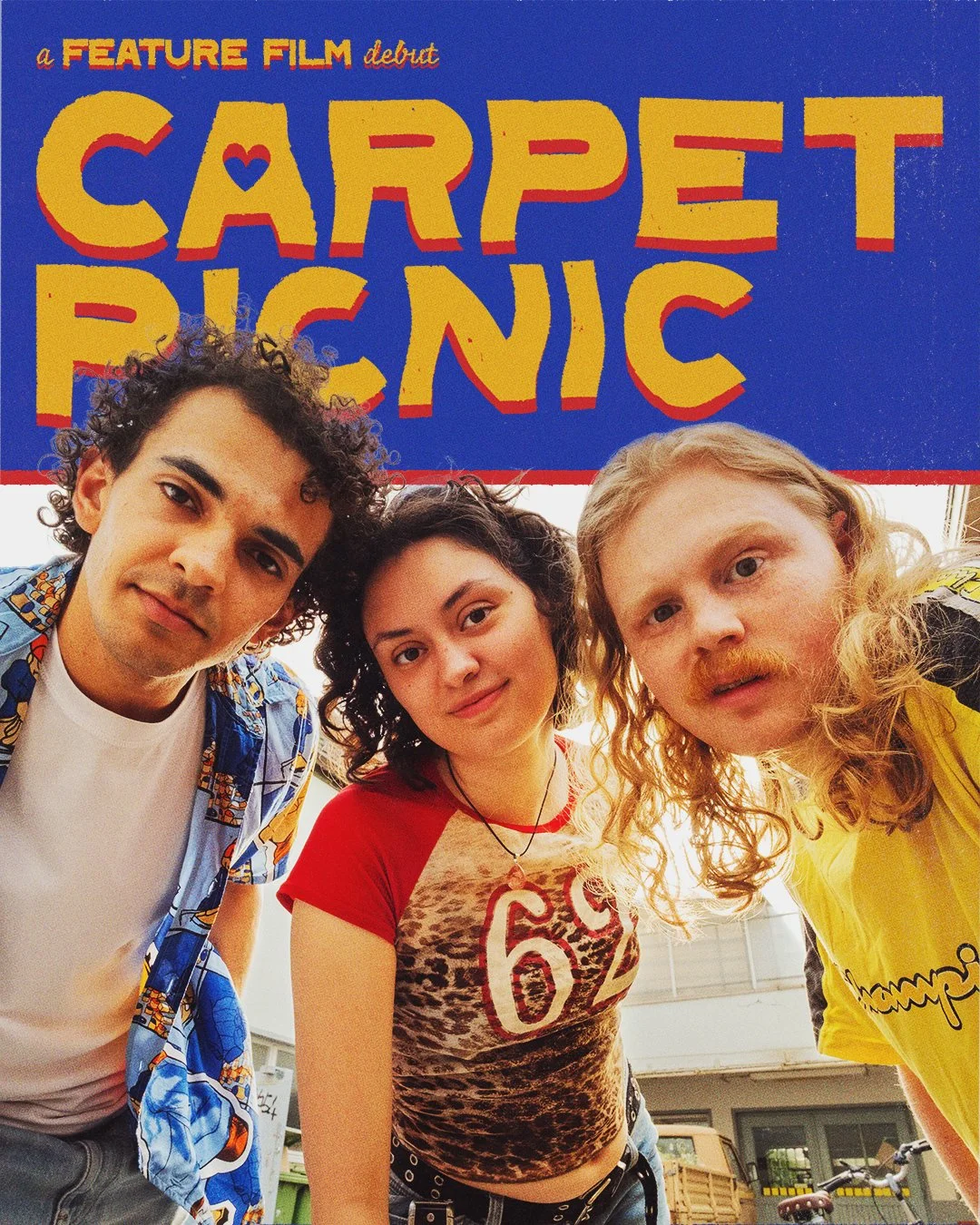 Three young people with curly and wavy hair looking at the camera, standing in front of a large sign that reads 'Carpet Picnic' with colorful, bold letters. The person on the left has dark curly hair and is wearing a patterned shirt over a white t-shirt. The person in the middle has dark wavy hair, wearing a red and animal print t-shirt. The person on the right has blonde curly hair and is wearing a yellow shirt.