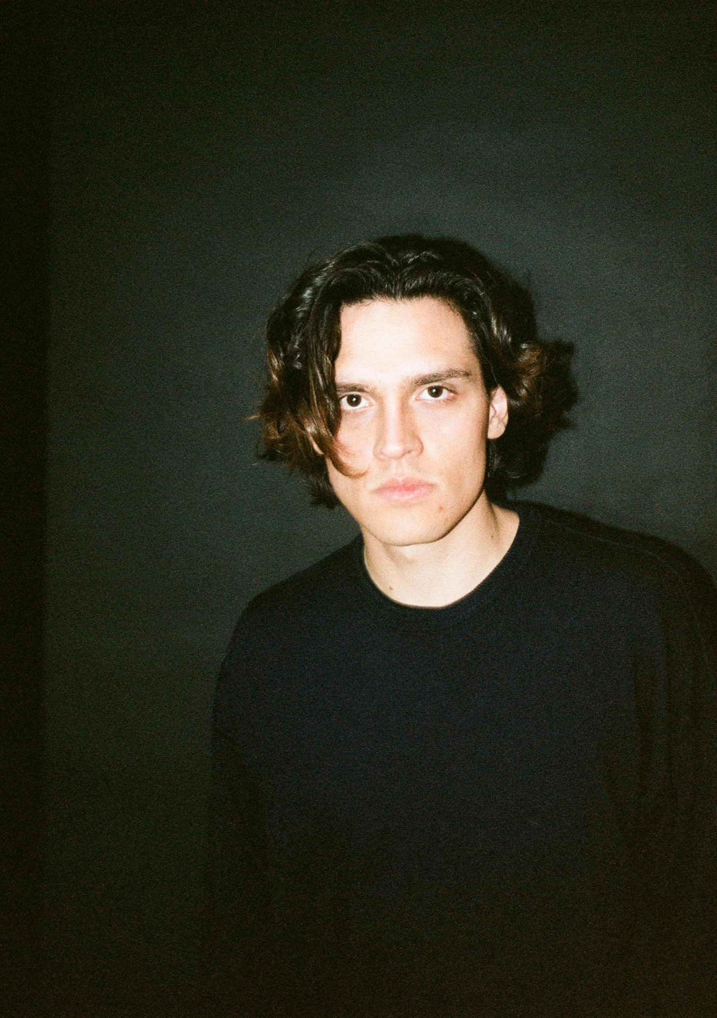 A young man with shoulder-length dark wavy hair and fair skin, wearing a black shirt, standing against a plain dark background.