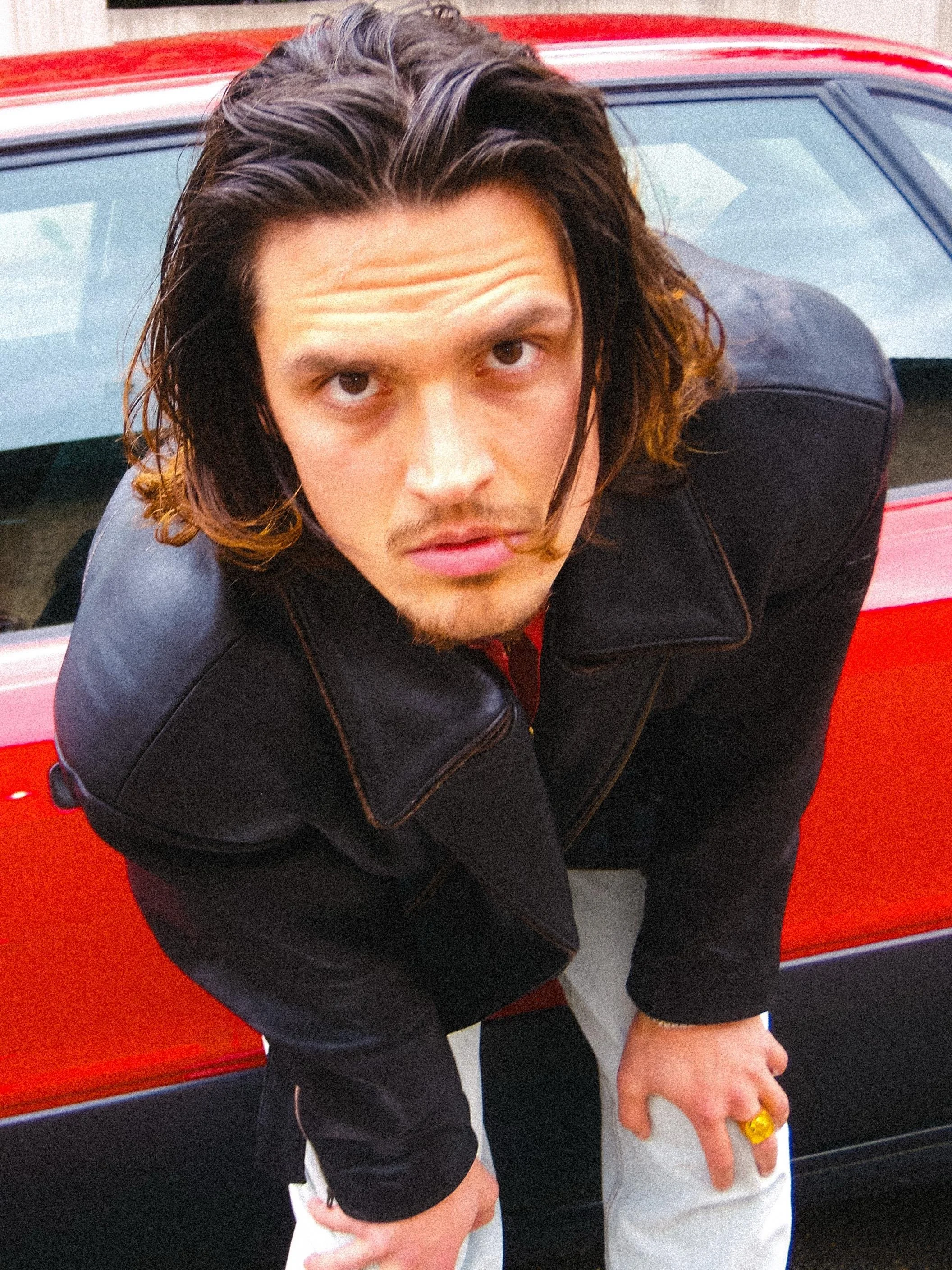 A person with long, dark, wavy hair leaning over a red car, wearing a black leather jacket and white pants with a gold ring on their right hand.