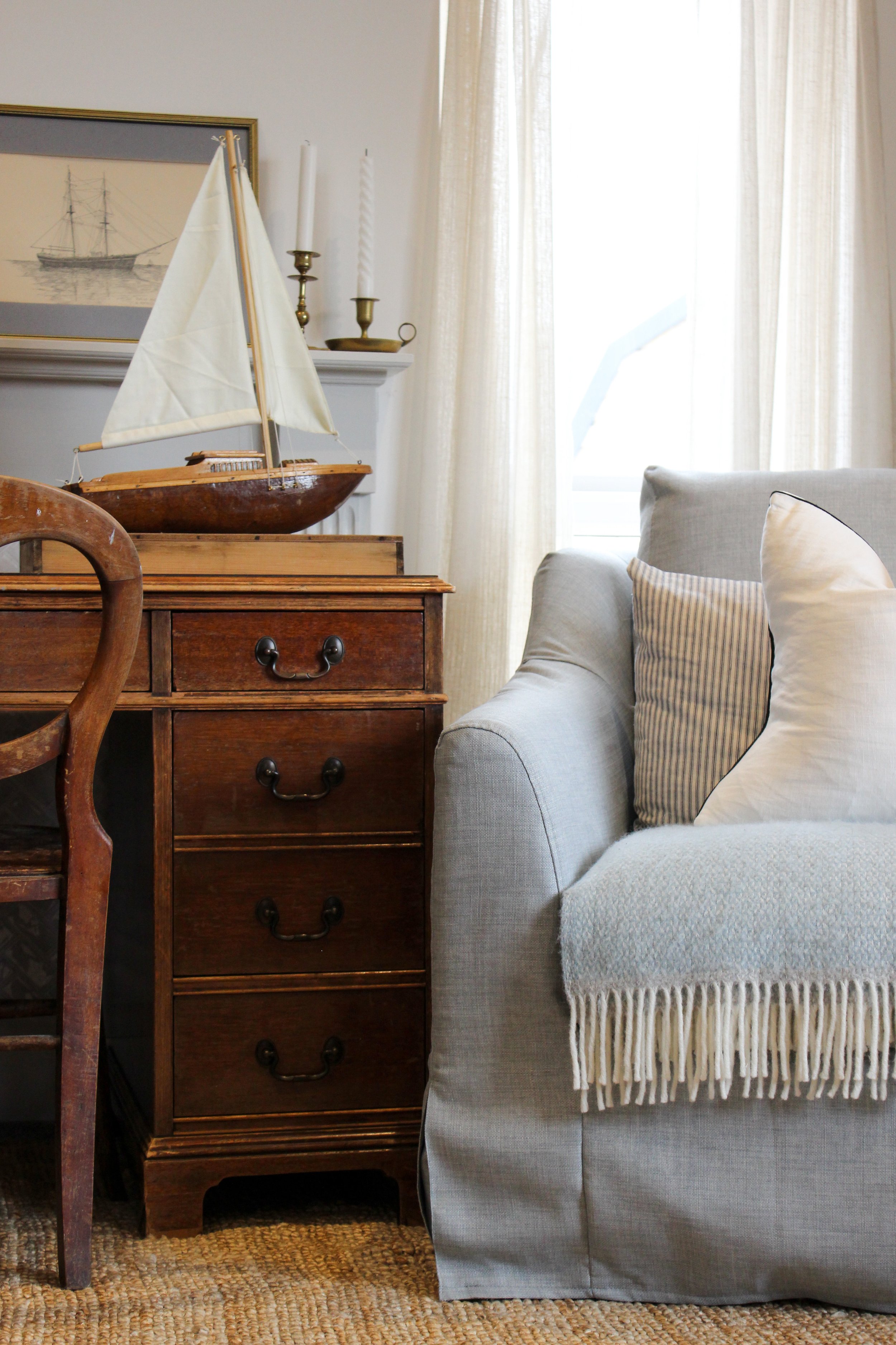A cozy living room corner with a wooden chest and a model sailboat on top, adjacent to a light gray upholstered sofa with pillows, next to a window with sheer curtains. A framed ship drawing and candlesticks adorn the background.