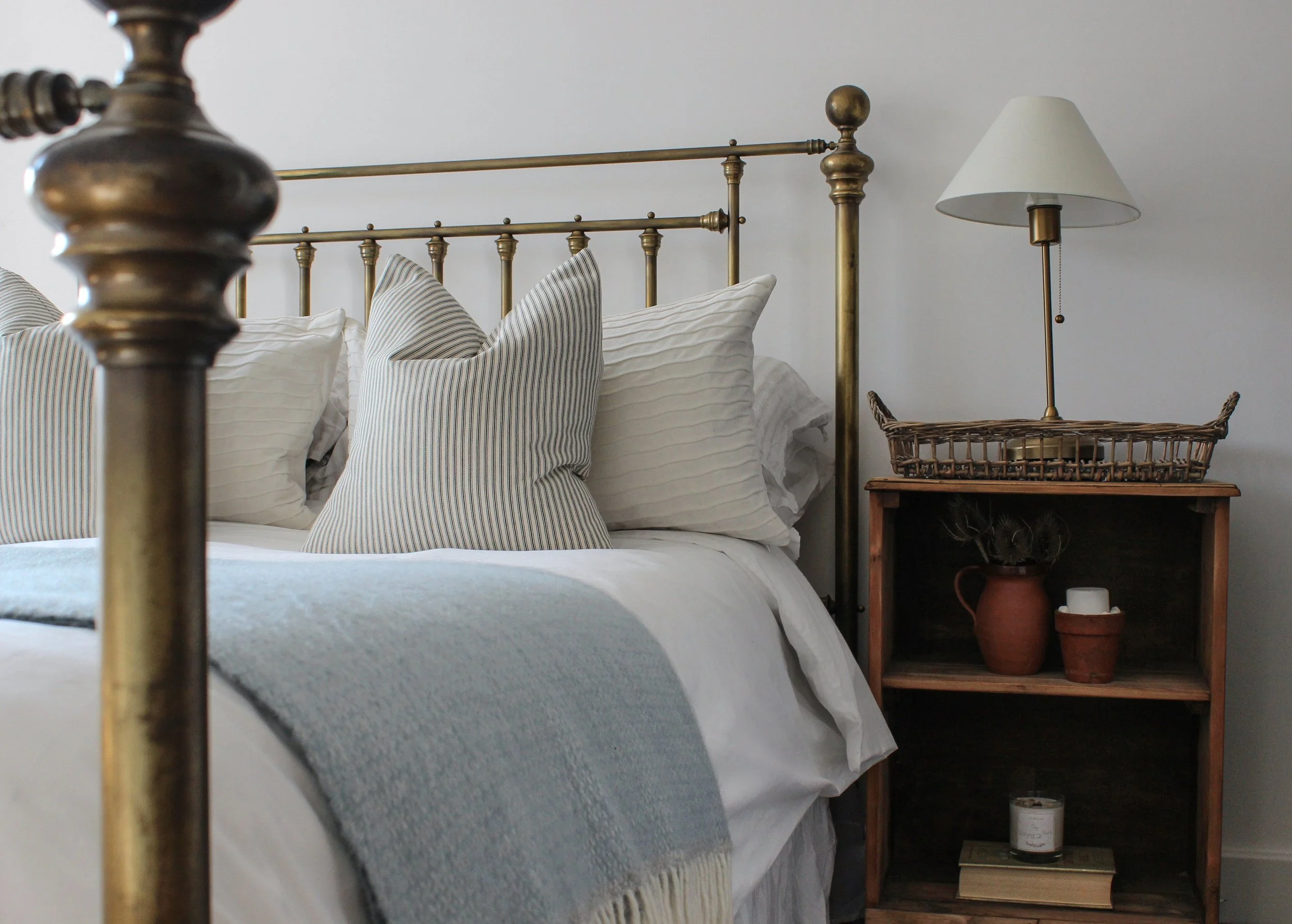 Close-up of a brass bed frame with white and striped pillows, beside a wooden nightstand with a white lamp, a large potted plant, and decorative candles, against a plain white wall.
