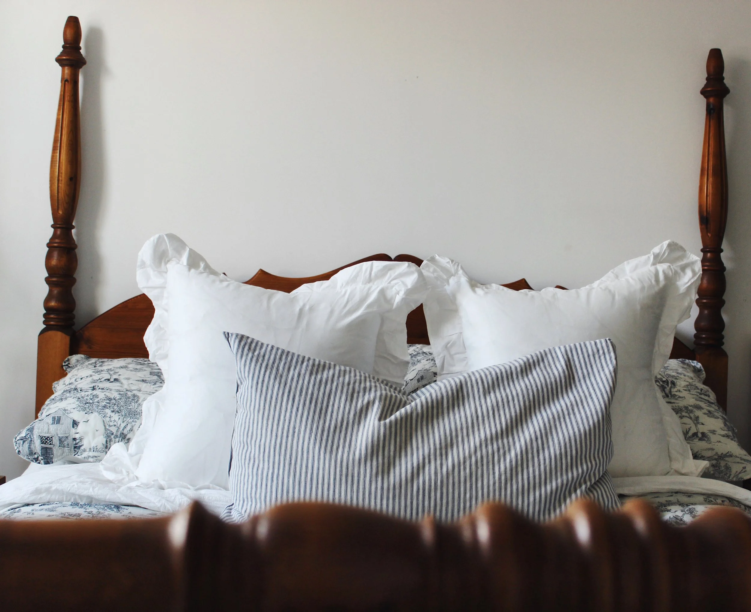 A bed with white and blue striped pillows, a white duvet, and a blue patterned bedspread. The headboard is wooden, and there are two wooden bedposts. The background is a plain off-white wall. Part of a person or animal's head, with brown fur, is partially visible at the bottom of the image.