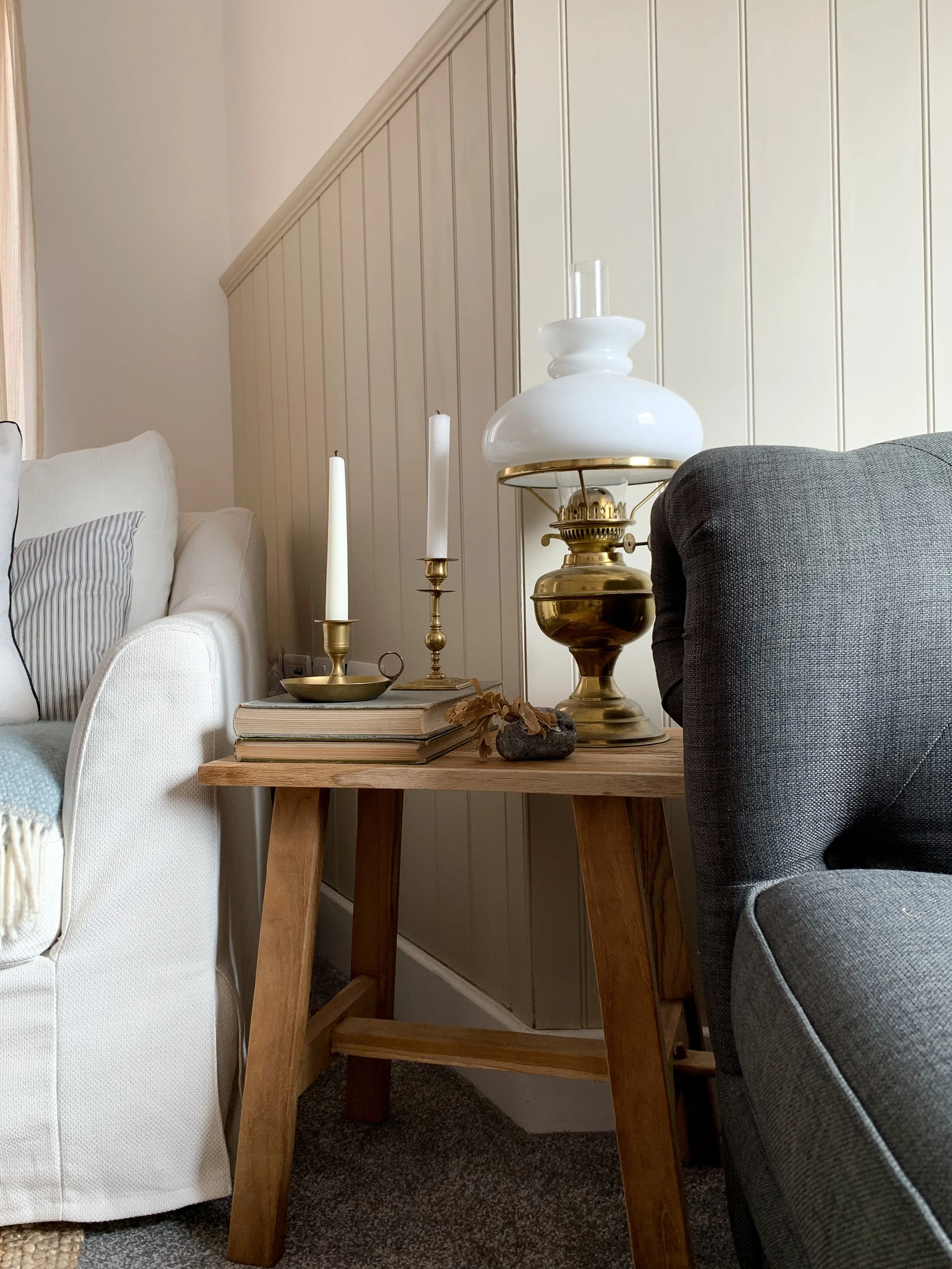 A living room corner with a white armchair, a gray upholstered chair, and a wooden side table with a vintage brass lamp, candles, books, and decorative stones.