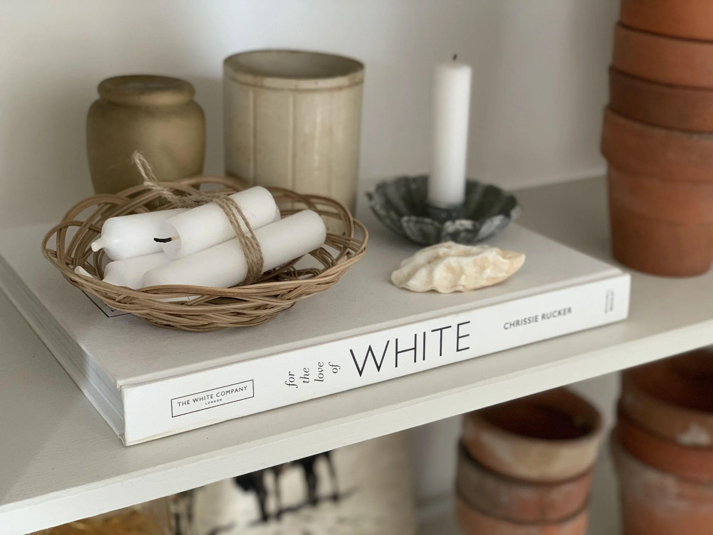 A display shelf holding a wicker basket with four white candles tied with string, two beige pottery vases, a white candle in a black and white ceramic holder, a rock, and a stack of terracotta pots.