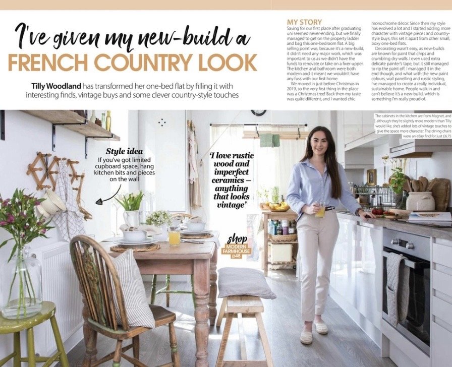 Magazine feature. A woman standing in a bright, modern kitchen with white cabinets and a wooden dining table, holding a glass of juice, surrounded by vintage-style decor and plants.