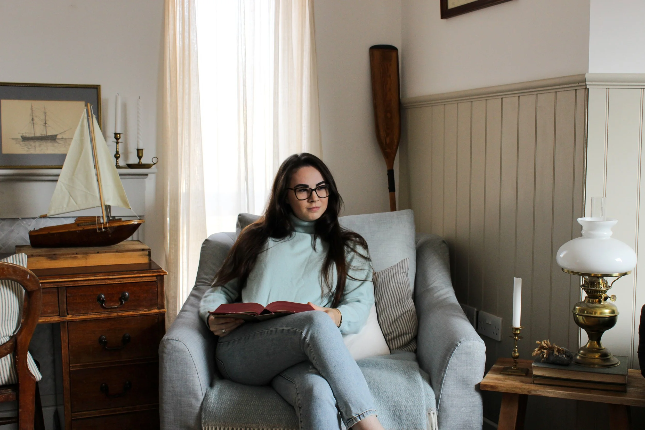 A woman with long dark hair and glasses sitting on a light gray sofa, reading a red book in a cozy living room with neutral-colored walls, a window with cream curtains, a wooden ship model, a framed picture of a sailing ship, a wooden chest of drawers, a paddle hanging on the wall, and a brass table lamp on a side table.
