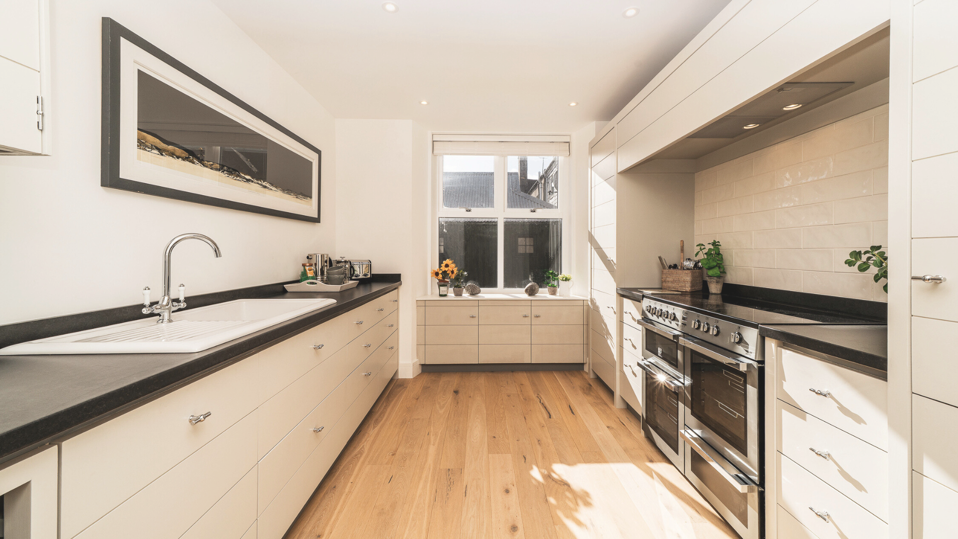 Bright kitchen with white cabinets, black countertops, and a large window with sunlight; includes a stove, plants, and decorative items.