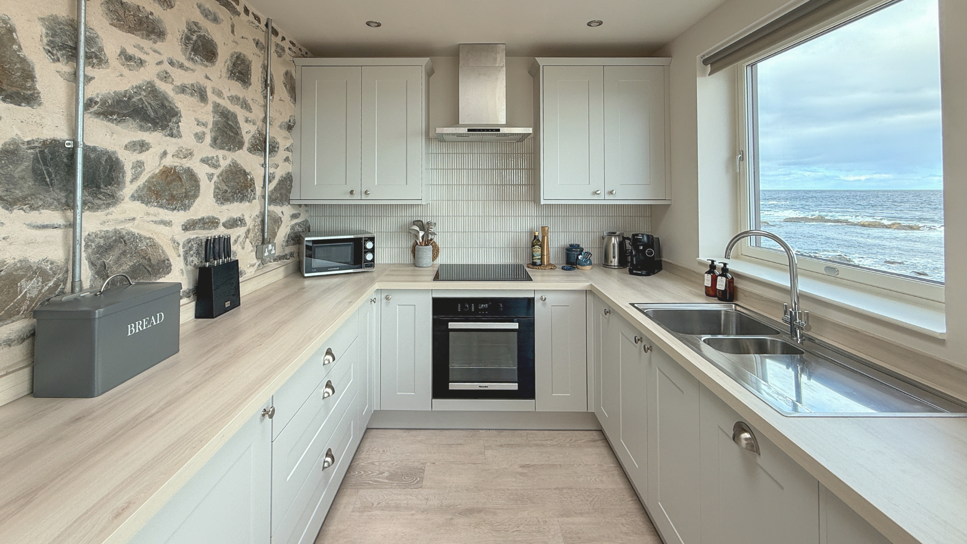 A kitchen with white cabinets, a stone accent wall, and a large window showing an ocean view. Countertops hold a bread box, microwave, utensils, and appliances.