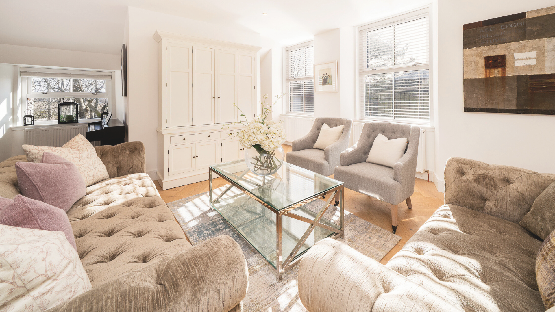 A cozy living room with beige and gray upholstered couches, pink and cream pillows, a glass coffee table with a floral centerpiece, and large windows with blinds allowing natural light.