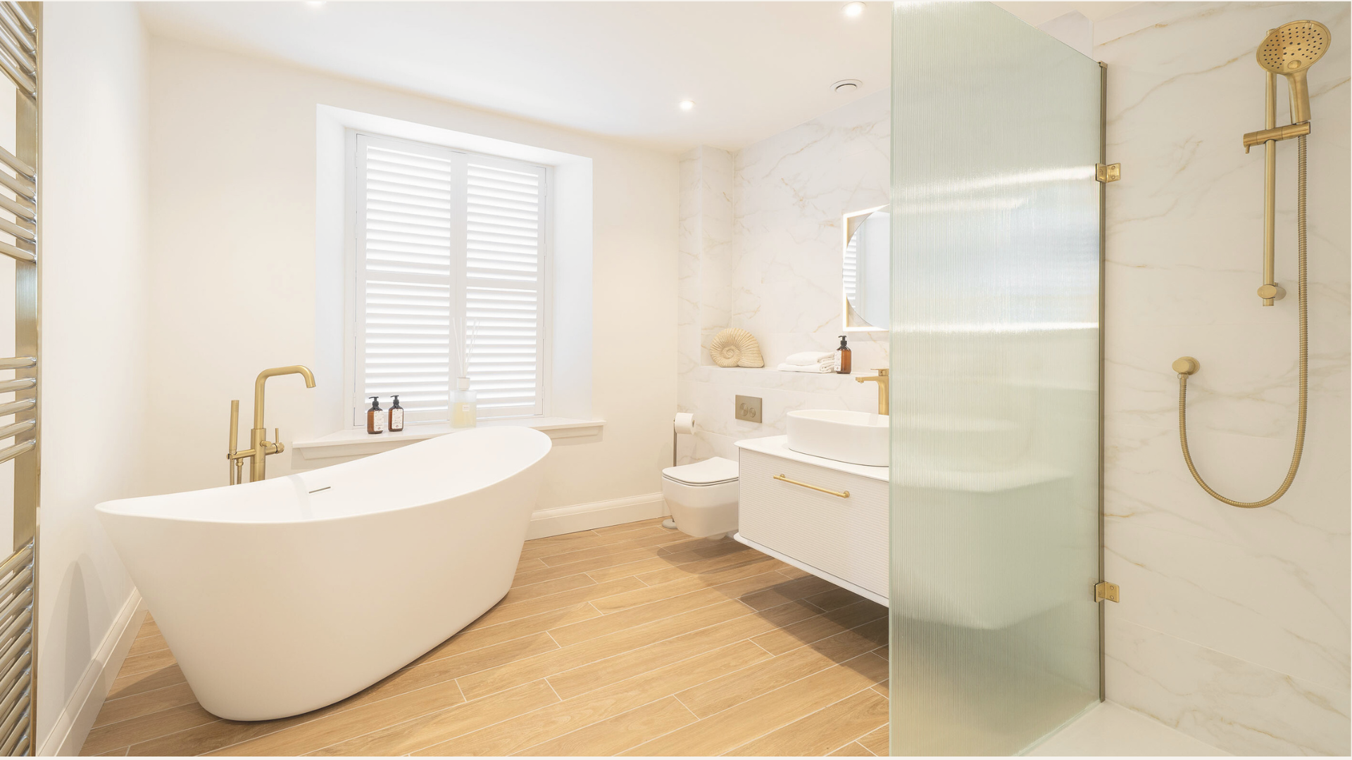 Modern, minimalist bathroom with a white free-standing bathtub, a window with shutters, a vanity with a vessel sink, a wall-mounted toilet, and a glass-enclosed shower with gold fixtures, marble walls, and wood-look floor tiles.