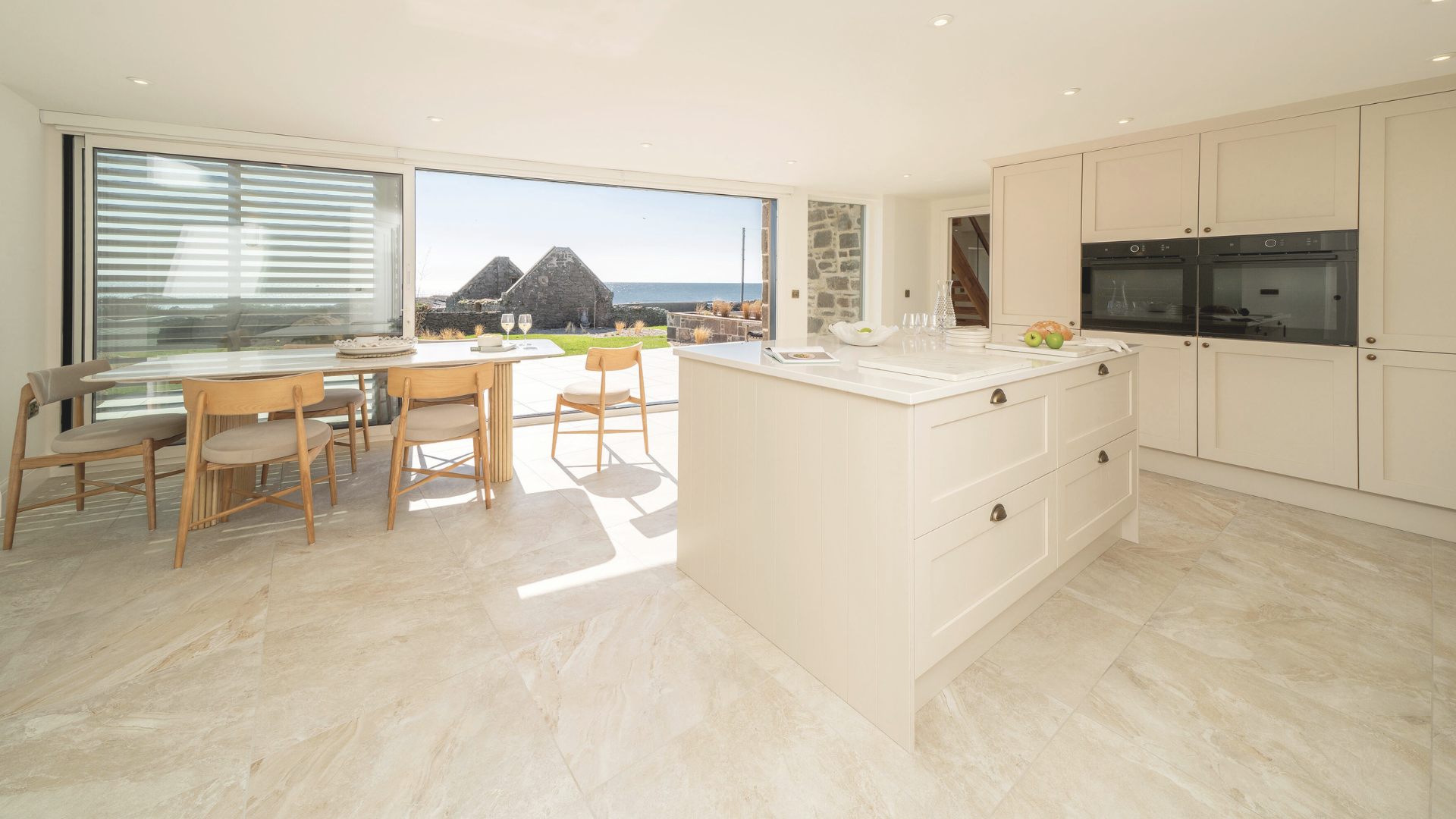 Bright modern kitchen with white cabinetry, an island with gray hardware, and a dining area with wooden chairs and a large sliding glass door overlooking a view of stone buildings, grass, and the sea.
