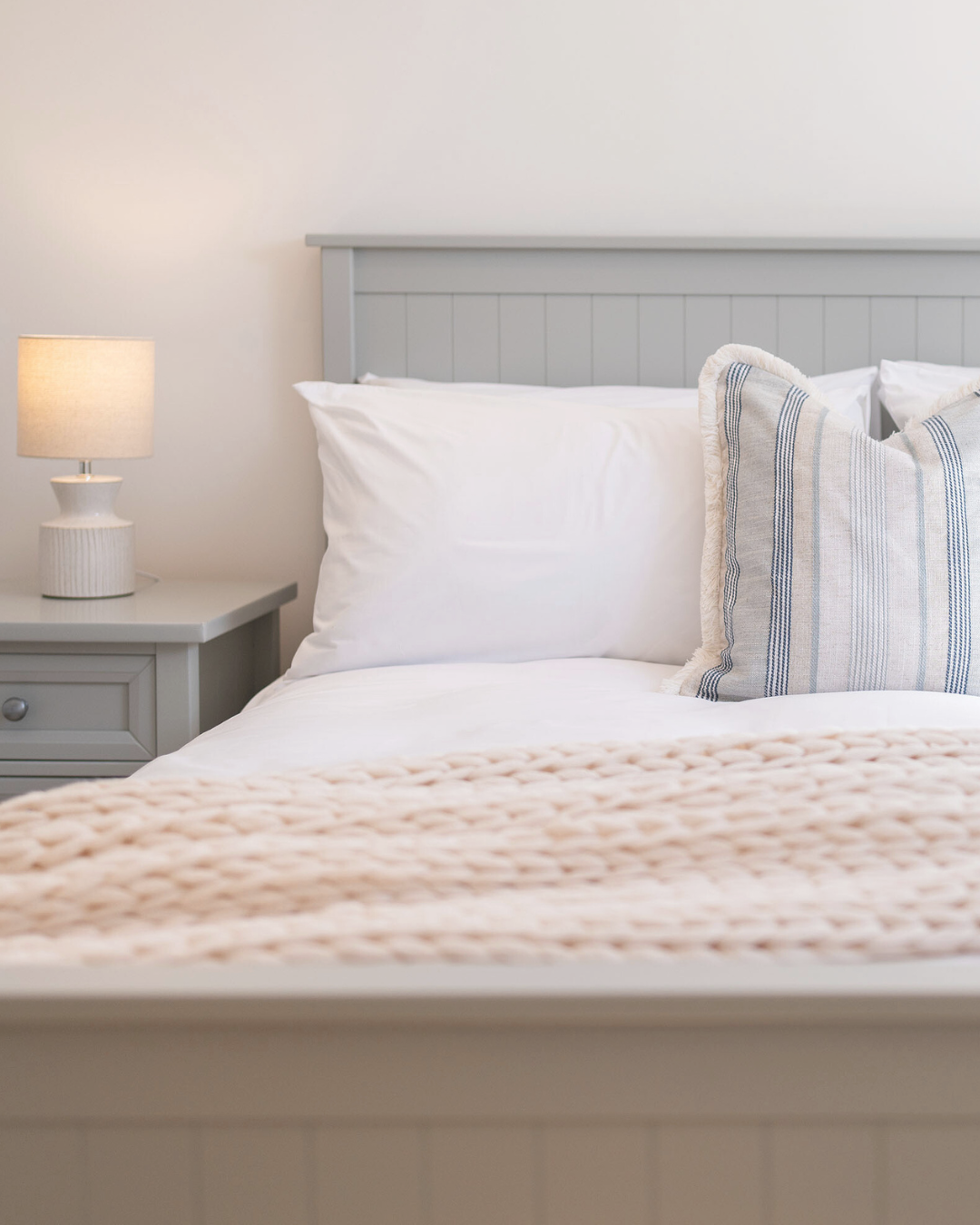 A neatly made bed with white pillows, a beige knitted blanket, and a striped pillow, beside a nightstand with a lamp in a minimalistic bedroom with white walls.