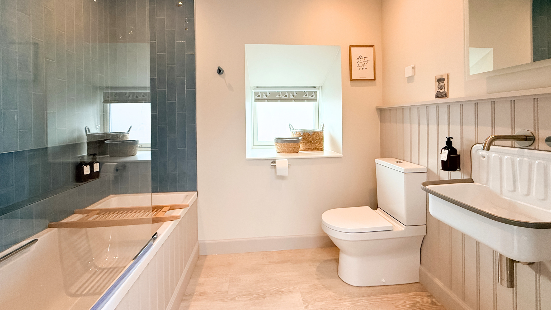 A bathroom with a bathtub on the left, a window with decorative curtains in the center, a toilet beneath the window, a small cabinet or storage unit above the toilet, a white sink with a brass faucet on the right, and a mirror above the sink. Decor includes framed art and a small picture on the wall.