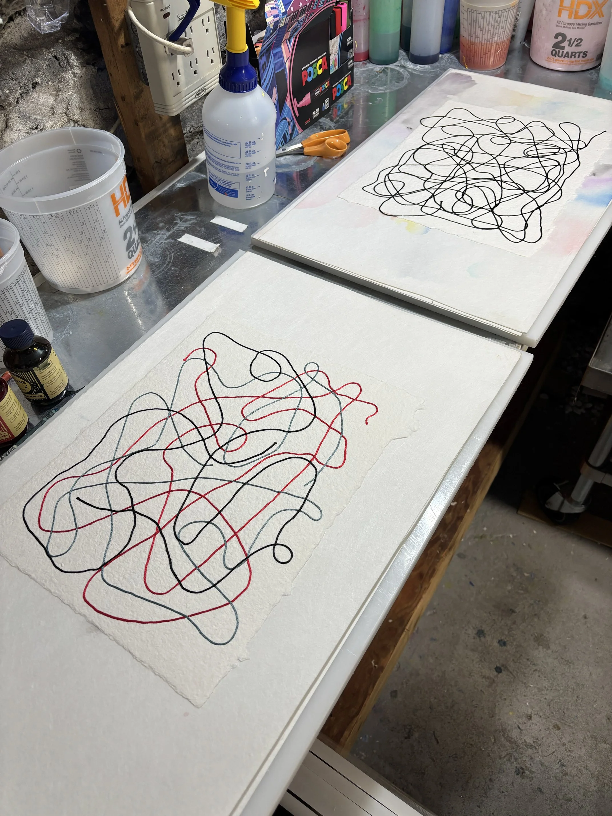 A view of the studio space with two newly created pieces of art, shown on a white workspace surrounded by art supplies including scissors, bottles, and containers.