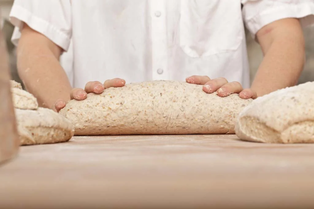 Person kneading dough on a wooden surface in a bakery kitchen.