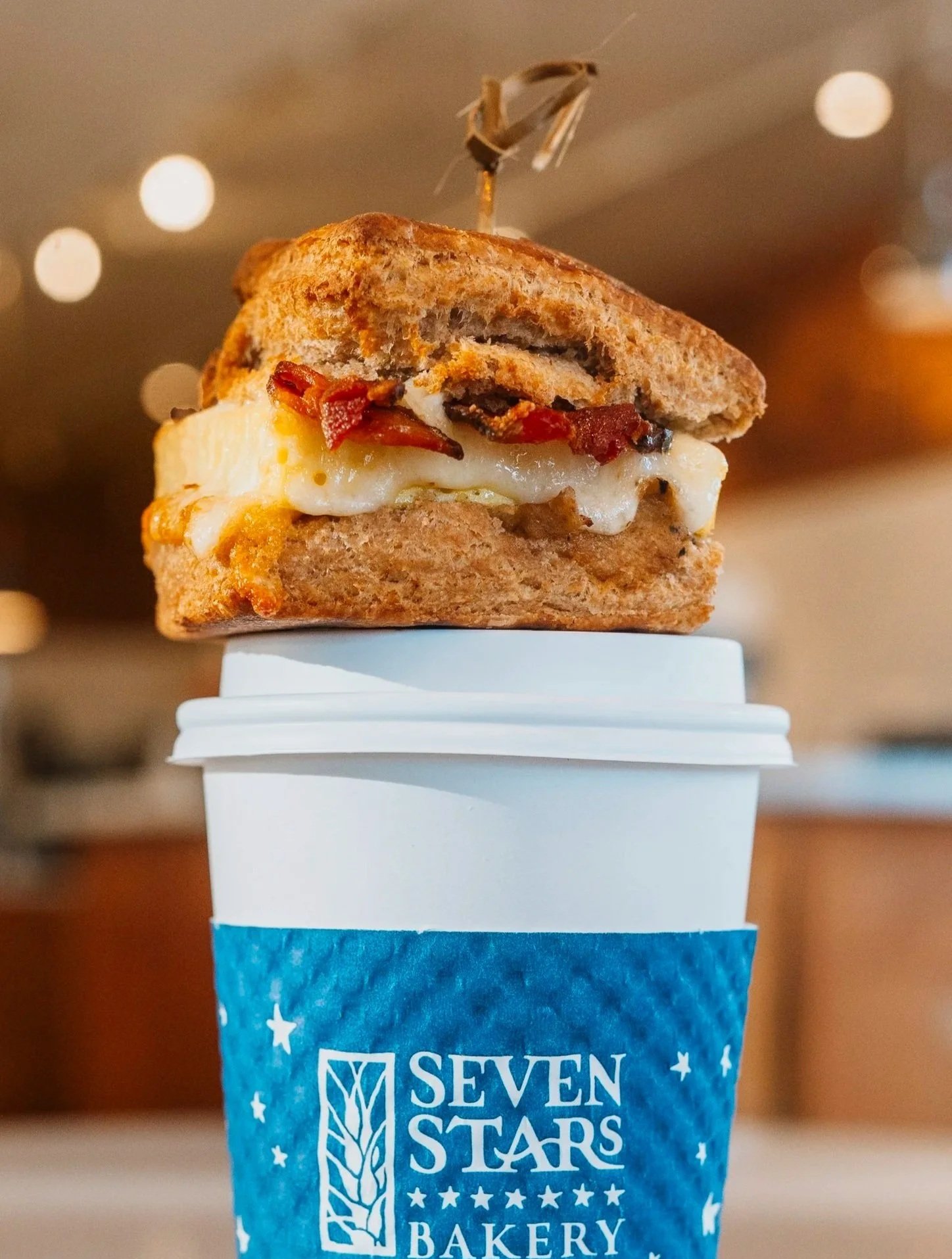 Close-up of a breakfast sandwich with a fried egg, bacon, and a biscuit in a blue paper cup labeled "Seven Stars Bakery" with blurred background lights.