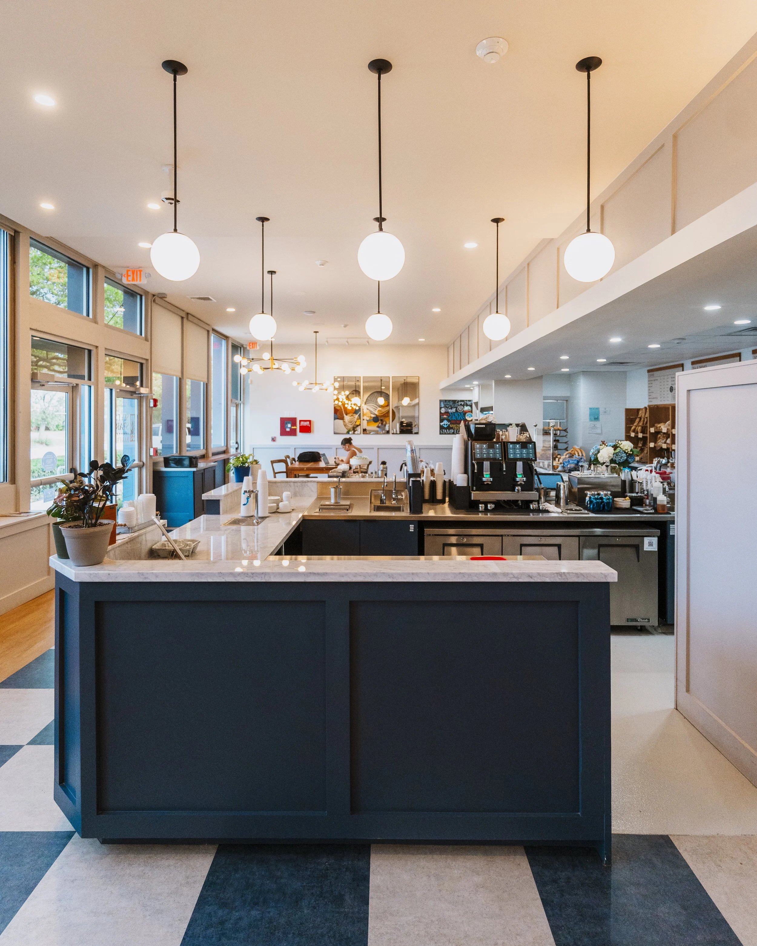 Interior of a modern cafe or coffee shop with a dark blue kitchen island counter, white marble top, potted plant, and a person sitting at a table in the background. Ceiling has round globe pendant lights, and large windows with trees outside.
