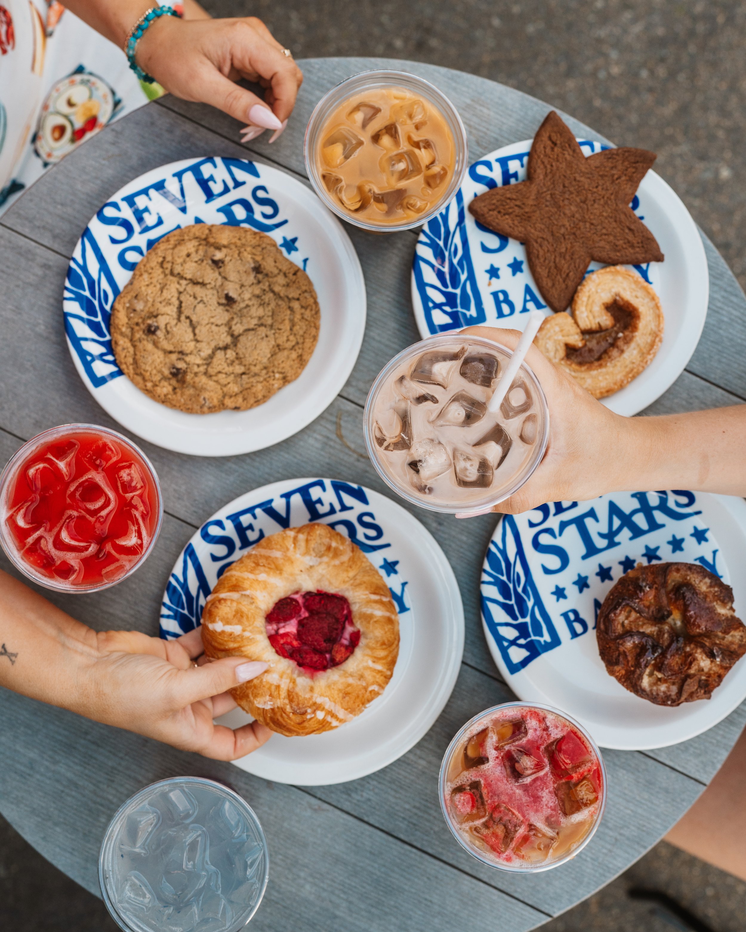 Overhead view of a croissant with red jam, iced coffees, cookies and pastries from Seven Stars Bakery on a wooden table.